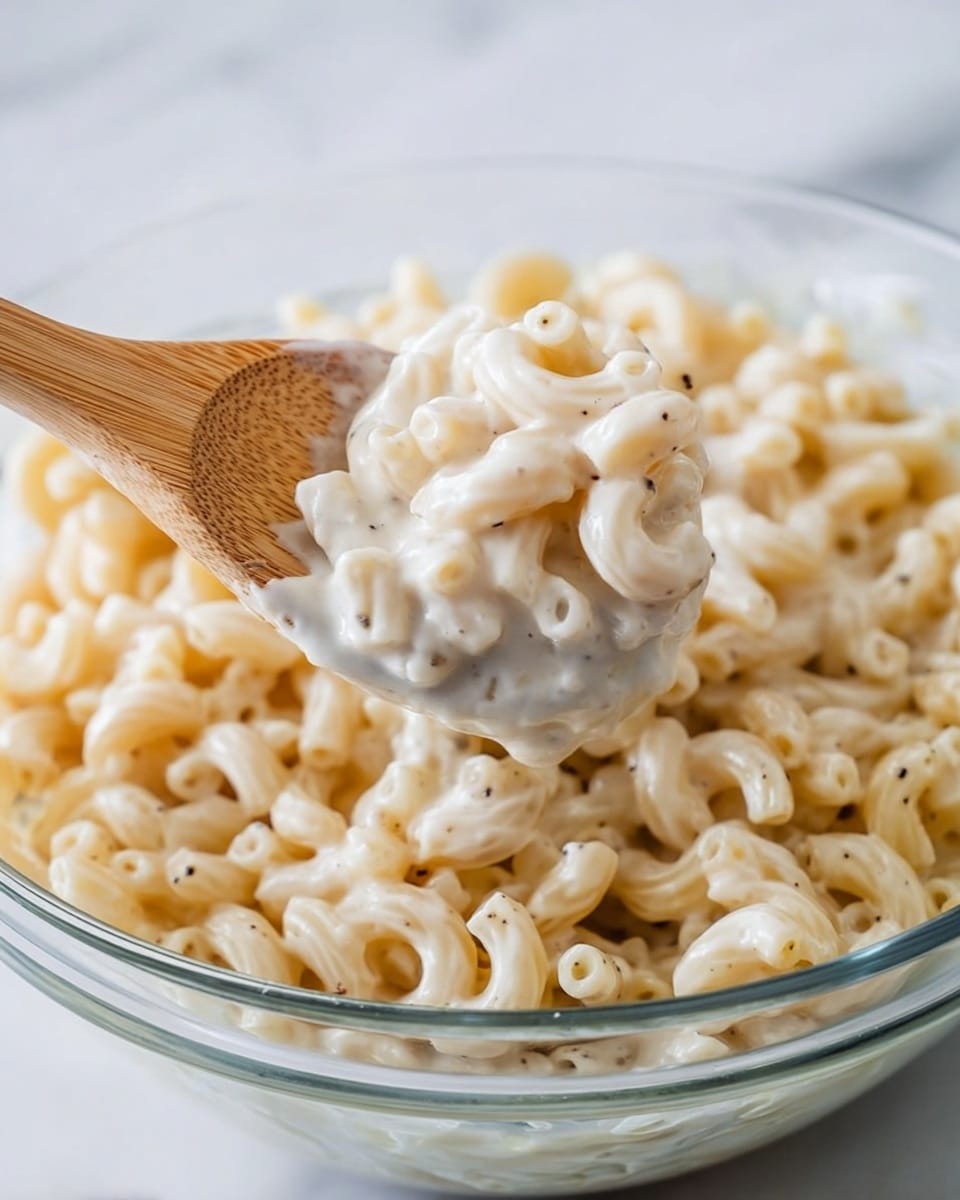 The image shows a clear glass bowl filled with cooked macaroni pasta, lightly coated with a creamy white sauce. A wooden spoon is lifting a portion of the macaroni, showing the smooth, thick sauce clinging to the pasta pieces. The macaroni is pale yellow with a soft texture, and the sauce looks rich and slightly speckled with black pepper. The bowl is placed on a white marbled surface. photo taken with an iphone --ar 4:5 --v 7