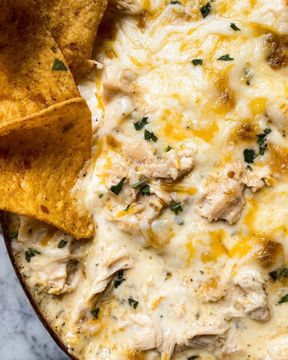 A close-up view of a creamy layered dish with melted white and yellow cheese on top, scattered chopped green herbs, and shredded pieces of light-colored poultry mixed throughout. At the bottom left, there are large light tan tortilla chips partially covered by the creamy, cheesy mixture. The entire dish rests on a surface with a white marbled texture. The melted cheese has a smooth and slightly stretchable look, with the creamy sauce giving a rich texture. Photo taken with an iphone --ar 4:5 --v 7