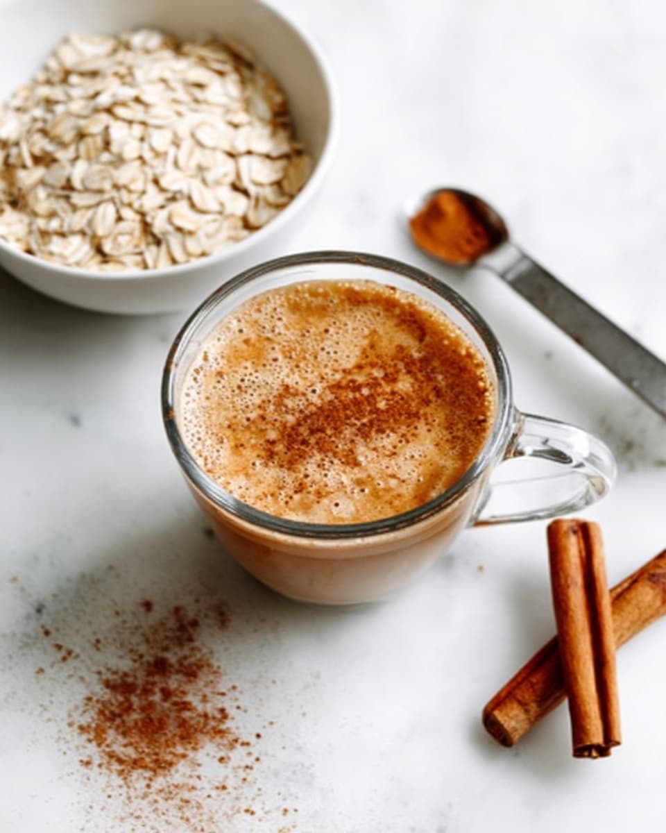 The image shows a clear glass cup filled with light brown creamy liquid sprinkled with a small amount of cinnamon powder on top. Below the cup, there is a white marbled surface. To the left side of the cup, there is a white bowl filled with rolled oats that look dry and pale. Two whole cinnamon sticks lie on the white marbled surface near the cup, adding a warm brown color to the scene. The overall setting is simple and clean, with soft natural light highlighting the textures. photo taken with an iphone --ar 4:5 --v 7