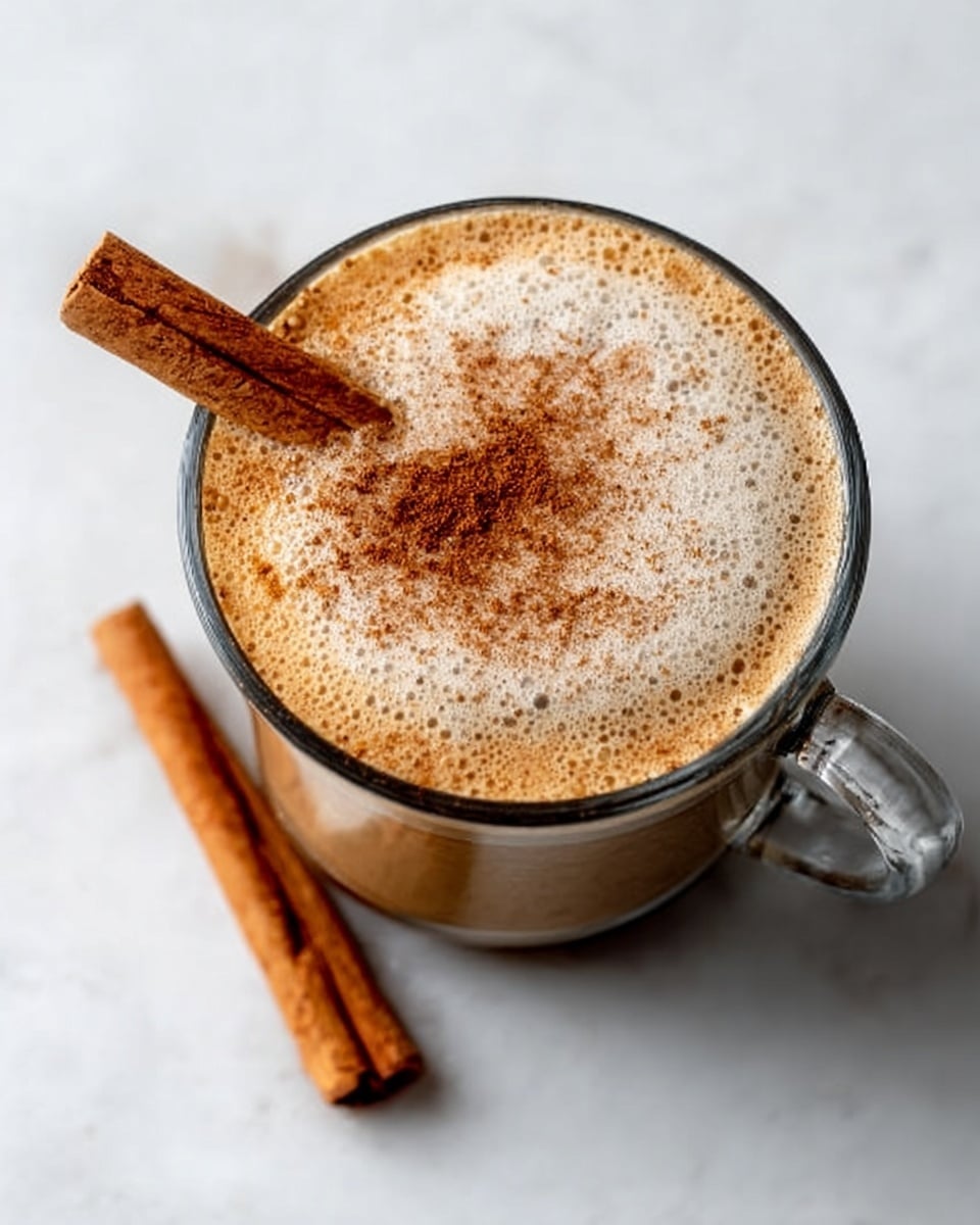 A clear glass cup filled with light brown creamy latte topped with frothy white foam sprinkled with cinnamon powder. Two cinnamon sticks rest on the side of the cup's rim, slightly overlapping. The cup sits on a white marbled surface with soft natural lighting. Photo taken with an iphone --ar 4:5 --v 7