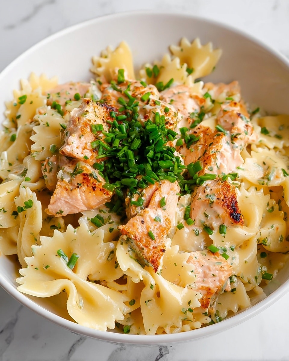 A white bowl filled with three main layers: the bottom layer has creamy, light yellow farfalle pasta with a smooth texture, then pieces of grilled pink salmon with some light char marks are mixed in, and the top layer is sprinkled with bright green chopped chives and some small green leafy herbs in the center, all placed on a white marbled surface. photo taken with an iphone --ar 4:5 --v 7