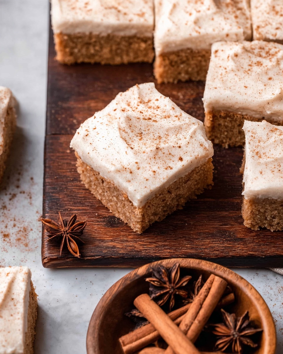 The image shows square pieces of spiced cake topped with a thick layer of light creamy frosting sprinkled with fine brown spice powder. The cake layer is light brown and looks soft and moist, while the frosting is smooth and covers the entire top evenly. The squares are arranged on a dark wooden surface, with some pieces slightly separated, showing the texture of the cake and frosting layer clearly. Next to the cake squares is a round wooden bowl filled with cinnamon sticks and star anise spices. The scene is set on a white marbled surface. photo taken with an iphone --ar 4:5 --v 7