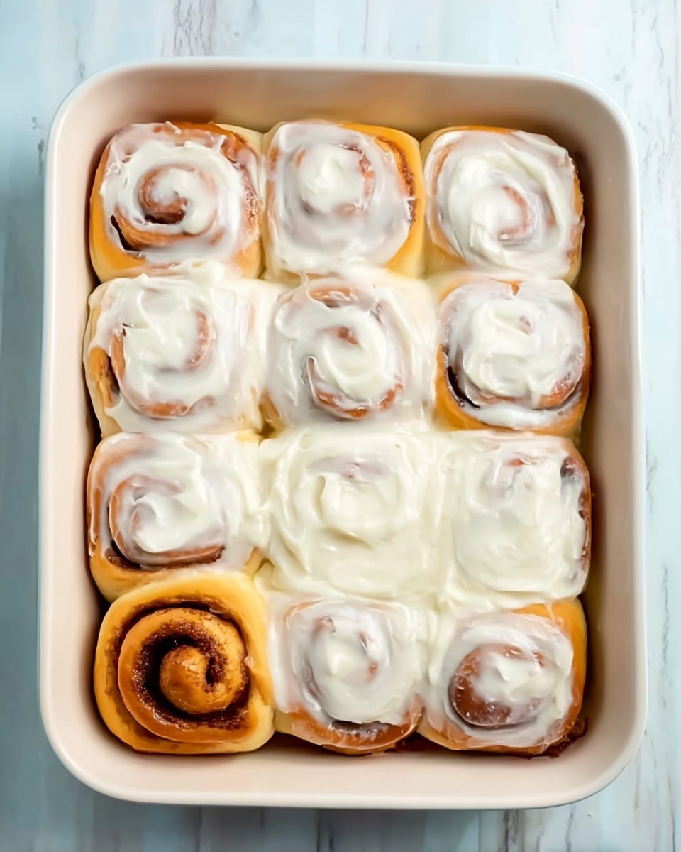 The image shows a white baking dish filled with nine cinnamon rolls arranged in three rows and three columns. Eight of the rolls are topped with thick white cream cheese frosting that covers most of the surface but still reveals swirls of the cinnamon filling underneath. One roll in the bottom left corner is plain, showing its golden-brown baked dough with dark cinnamon swirl lines clearly visible. The dish is set on a white marbled surface, and the texture of the rolls looks soft and fluffy with smooth creamy frosting. photo taken with an iphone --ar 4:5 --v 7