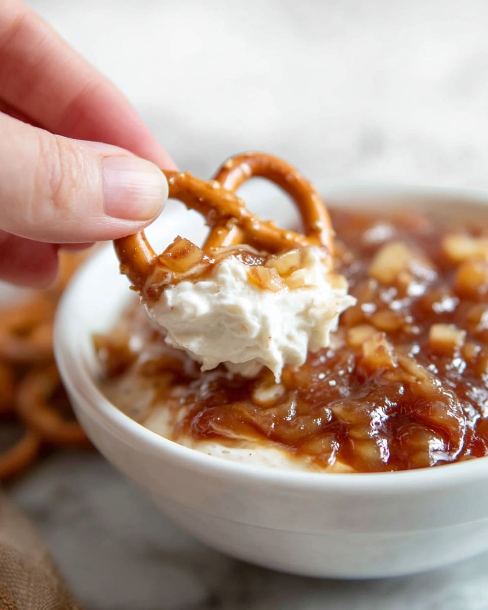 A close-up of a woman's hand holding a small brown pretzel topped with a creamy white mixture and small chunks of caramel-colored fruit, with a white bowl underneath filled with two layers: a bottom layer of the creamy white mixture and a top layer of chunky caramel fruit glaze that looks glossy and syrupy, all placed on a white marbled surface. photo taken with an iphone --ar 4:5 --v 7