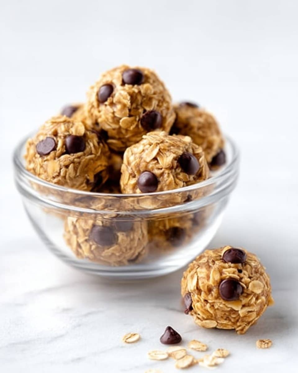 The image shows a clear white glass bowl filled with round, light brown oat cookies topped with dark chocolate chips. Each cookie is rough in texture, showing visible oats pressed together with some chocolate chips placed on top. One cookie sits outside the bowl on the right side on a white marbled surface. The overall look is simple, with the cookies stacked inside the bowl and the background plain white. photo taken with an iphone --ar 4:5 --v 7