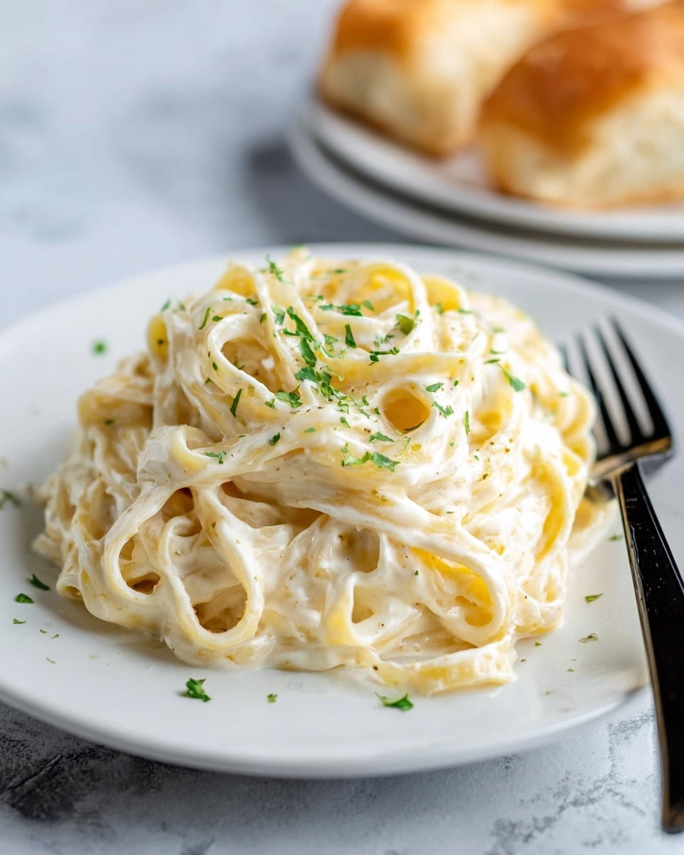 The image shows a white plate with a pile of creamy fettuccine pasta covered in a smooth, white Alfredo sauce. The pasta strands are thick and twisted neatly in a mound at the center of the plate. Small green parsley flakes are sprinkled lightly over the top, adding color contrast. The plate is placed on a white marbled surface. In the background, blurry pieces of bread sit on another white plate, and a fork with a black handle is resting partially under the pasta on the right side. photo taken with an iphone --ar 4:5 --v 7