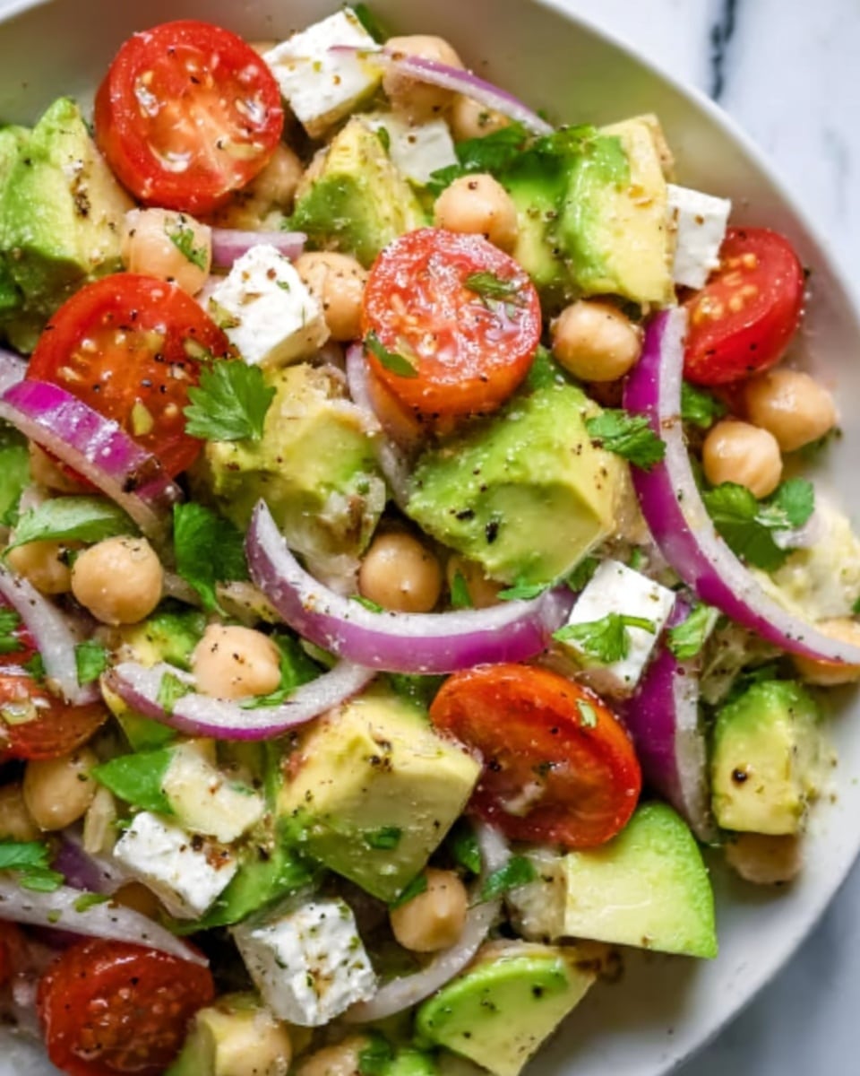 The image shows a fresh salad with several layers of ingredients visible. The base layer has light green avocado pieces with a smooth texture, mixed evenly with cream-colored chickpeas that are round and firm. Bright red cherry tomato halves, juicy and shiny, are scattered across the salad along with white cubes of feta cheese that have a slightly crumbly texture. Thin slices of purple onion add contrast, spread throughout the dish, and small green leaves of cilantro or parsley are sprinkled on top. The salad is on a white plate, sitting on a white marbled surface. The photo taken with an iphone --ar 4:5 --v 7