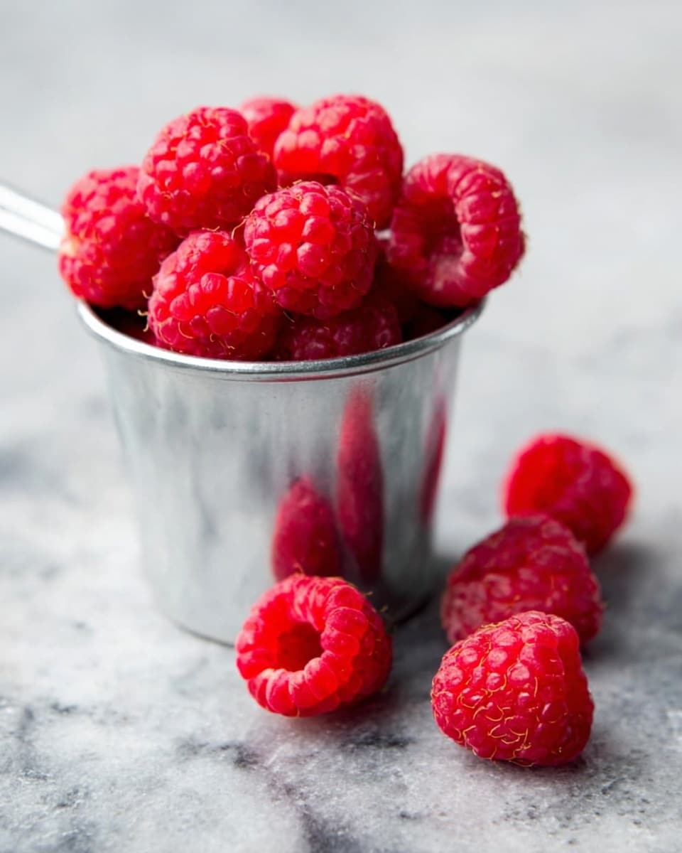 A small metal cup filled with fresh bright red raspberries, piled high and slightly overflowing, sits on a white marbled surface. Next to the cup are six scattered raspberries, showing their bumpy, soft texture and vibrant red color. The metal cup has a simple, smooth texture and reflects some light softly. The background is blurred but maintains the white marbled texture, making the raspberries stand out clearly. photo taken with an iphone --ar 4:5 --v 7