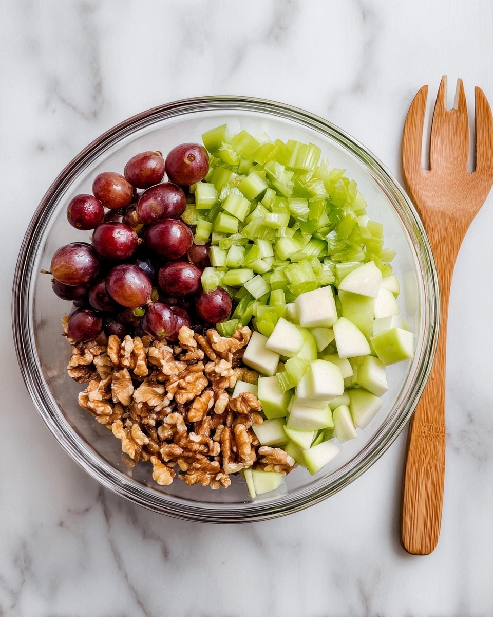 A clear glass bowl holds four separate layers of salad ingredients arranged side by side: dark red grape halves on the upper left, light green chopped celery on the upper right, light green apple cubes with white flesh on the lower left, and a pile of light brown walnut halves on the lower right. The bowl is placed on a white marbled surface. To the right of the bowl, there is a wooden salad fork lying flat. photo taken with an iphone --ar 4:5 --v 7