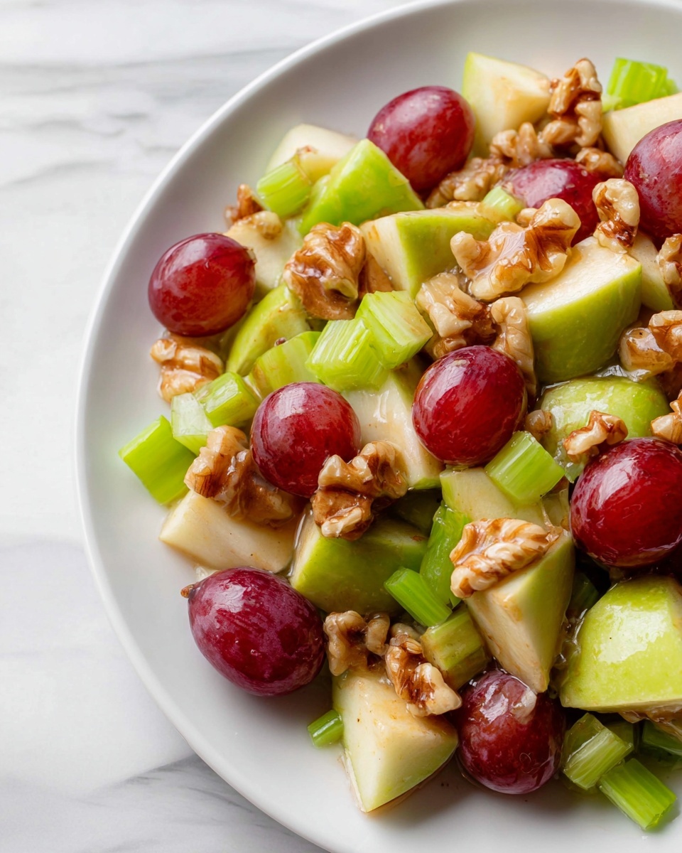 The image shows a close-up of a white plate with a fresh fruit and nut salad. The salad has three main layers: large chunks of green apple with bright green skin, whole and halved red grapes with a shiny surface, and light brown walnut halves scattered throughout. Small bright green celery pieces are mixed in, adding extra texture. The ingredients have a glossy coating that makes them look fresh and juicy. The plate is set on a white marbled surface. Photo taken with an iphone --ar 4:5 --v 7