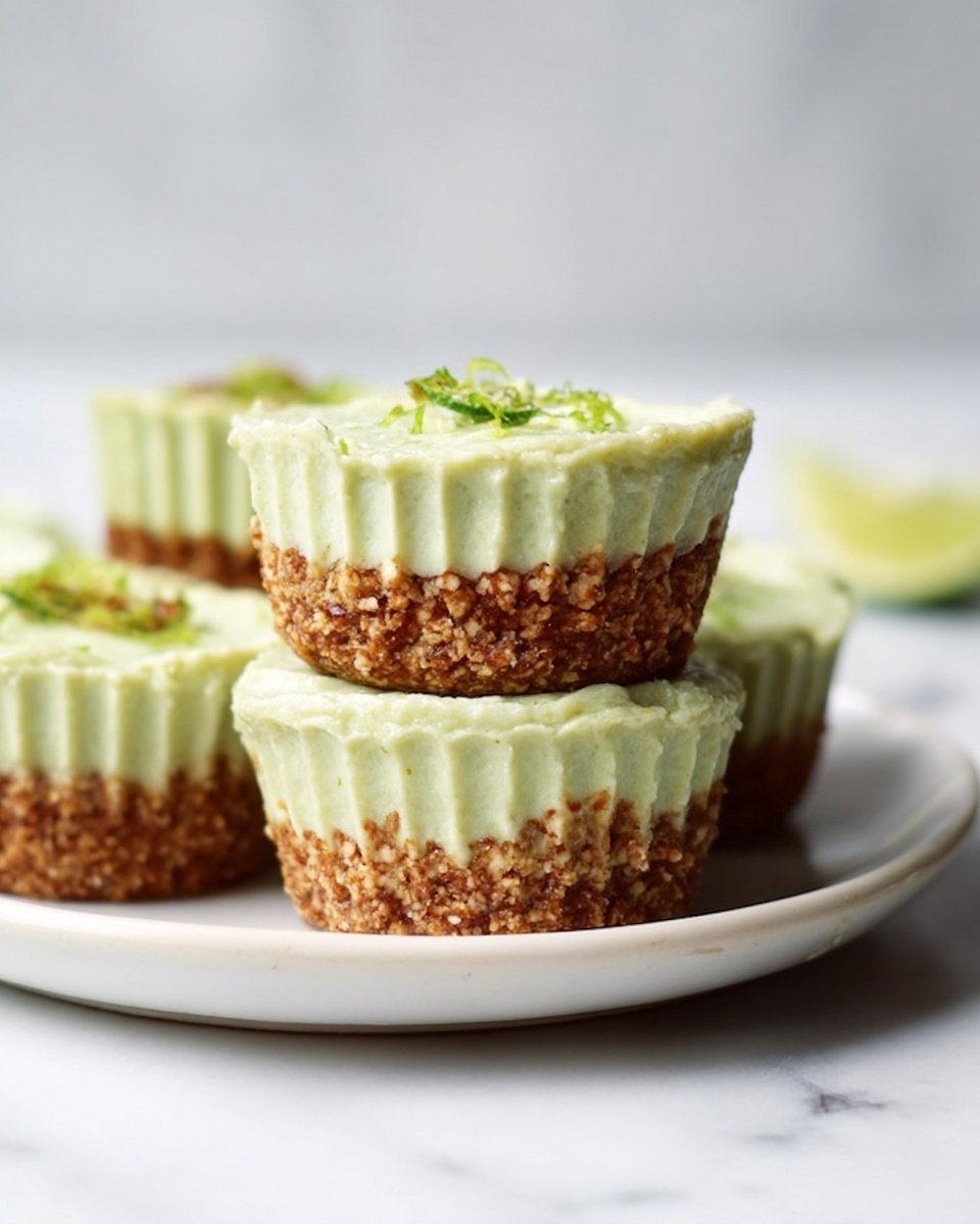 The image shows a close-up of three small dessert cups stacked on a white plate, set on a white marbled surface. Each cup has two clear layers: the bottom layer is a crumbly light brown crust made of nuts or grains with a coarse texture, while the top layer is a smooth, thick light green cream that appears soft and fluffy, covering the crust. The cream layer has small ridges along the sides, showing it was pressed into cupcake liners. One cup features a small piece of lime and some green zest on top, adding a fresh look. In the background, there are more dessert cups slightly blurred. Photo taken with an iphone --ar 4:5 --v 7