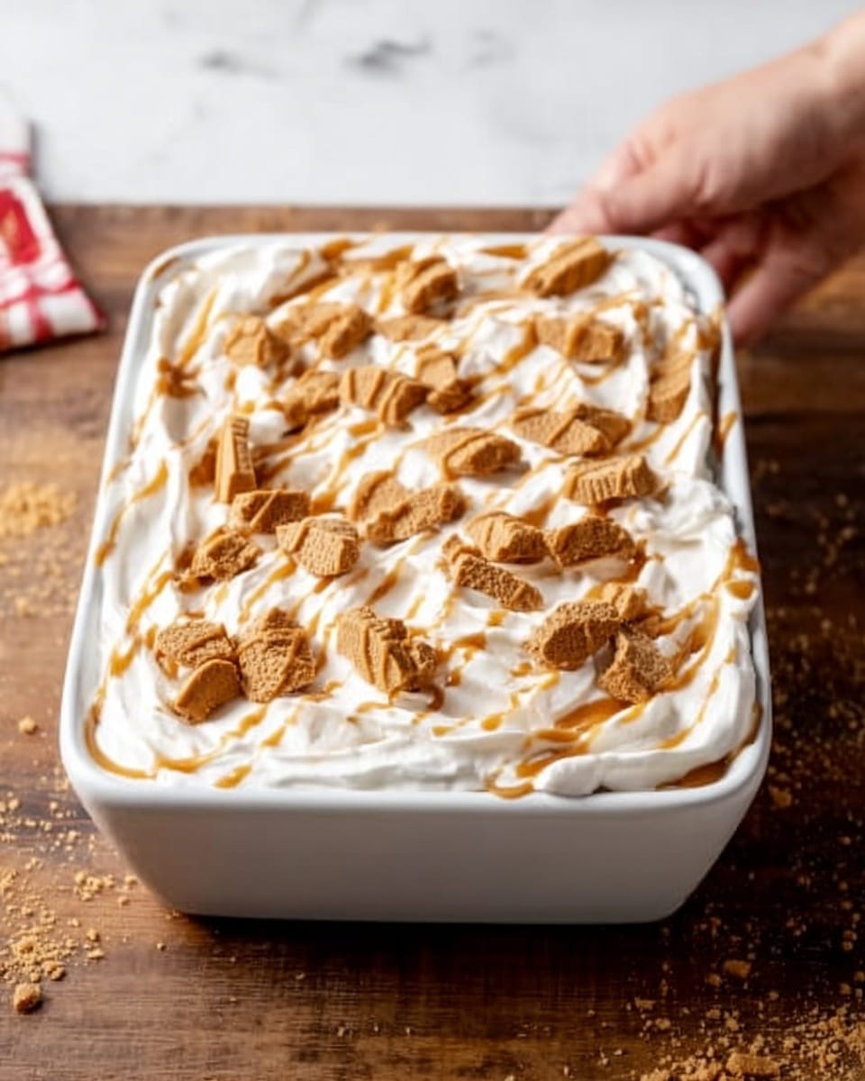 The image shows a rectangular white ceramic dish filled with a dessert topped with a thick layer of white whipped cream. Over the cream, there is a light brown sauce drizzled in a swirling pattern. Crushed light brown cookie pieces are evenly scattered on top, adding texture and color contrast. The dish is placed on a wooden surface with a white marbled texture background visible around it. A woman's hand is reaching toward the dish from the left side. Photo taken with an iphone --ar 4:5 --v 7