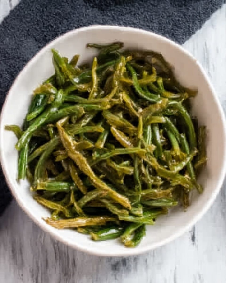 A white bowl filled with cooked green beans that look soft and slightly shiny, arranged loosely inside the bowl. The green beans have a darker green color with some light brown spots, showing they are cooked well. The bowl is placed on a white marbled surface with a dark cloth partially under it on one side, creating a simple and clean setting. Photo taken with an iphone --ar 4:5 --v 7