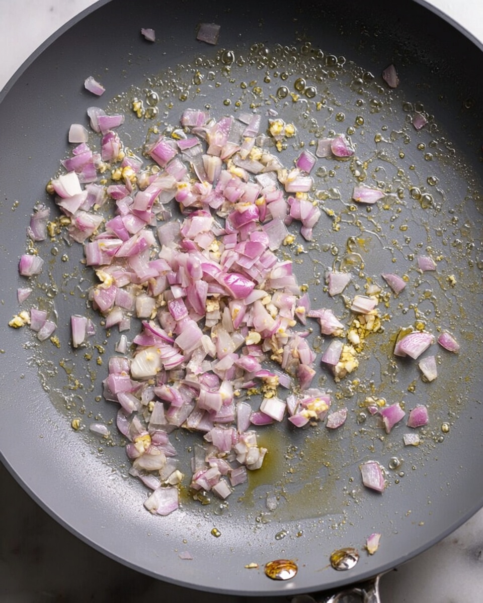 A close-up top view of a gray frying pan with small chopped pieces of light purple and white shallots and tiny bits of minced garlic sizzling in bubbling oil, scattered unevenly across the pan surface. The pan rests on a white marbled background with its metal handle partially visible on the right side. The light reflects softly on the oil, showing a thin layer coating the pan photo taken with an iphone --ar 4:5 --v 7