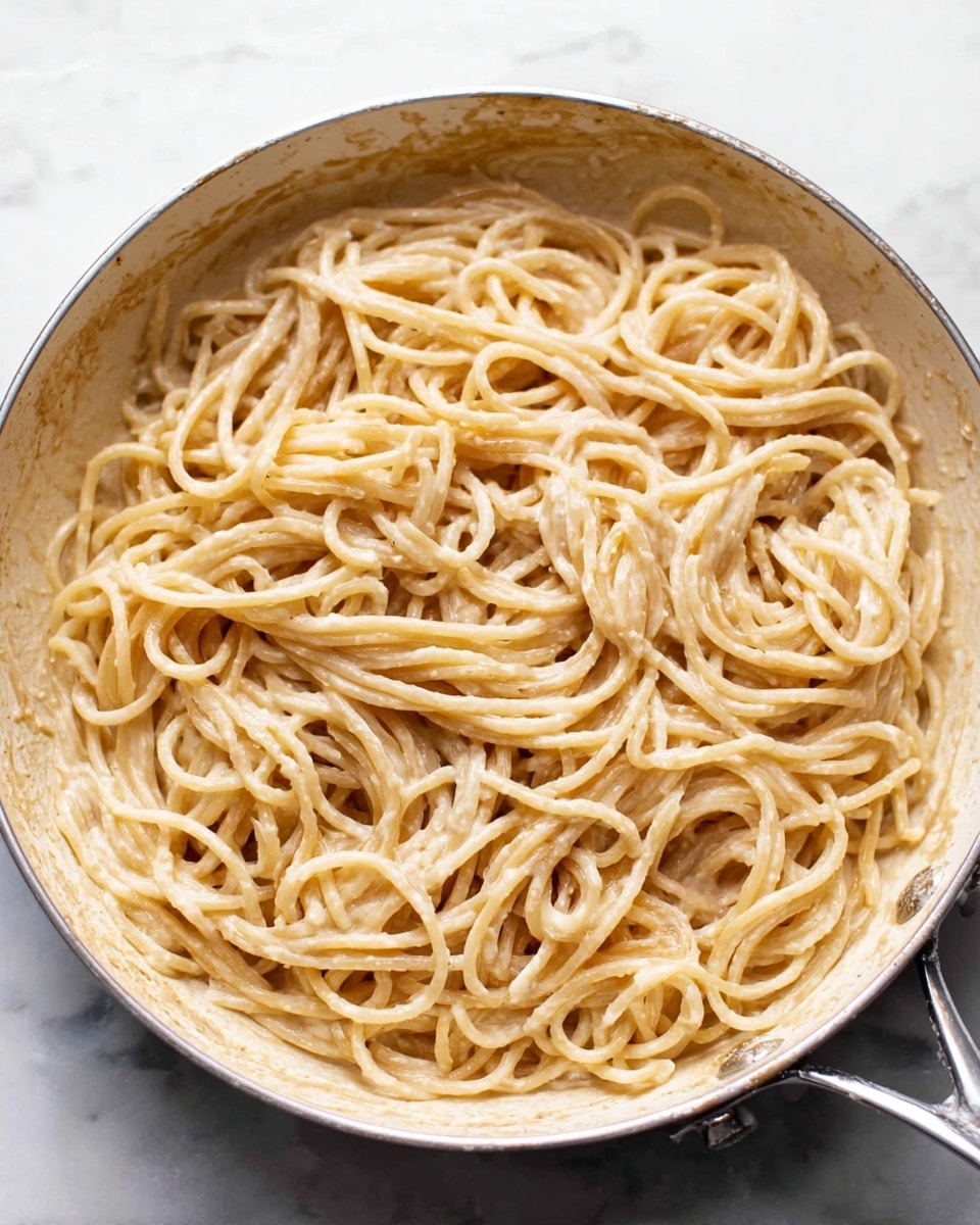 A white pan filled with long, round spaghetti noodles that are coated in a creamy, light beige sauce. The spaghetti is well mixed with the sauce, showing a smooth texture with some sauce pooled at the bottom. The edges of the pan have a light coating of sauce residue, and the pan is placed on a white marbled surface. photo taken with an iphone --ar 4:5 --v 7