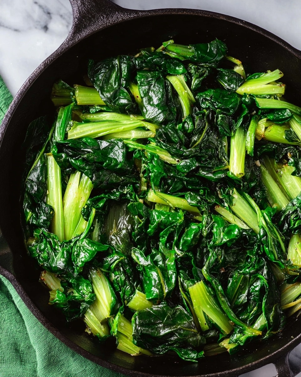 This image shows cooked leafy greens in a black cast iron pan. The dish has two layers: the first layer is the light green, thick stalks of the leafy vegetable, and the second layer is the dark green, wilted leafy parts that cover most of the pan. The greens look moist and glossy, showing they are cooked but still fresh. The pan sits on a white marbled surface with part of a green cloth visible at the bottom left. Photo taken with an iphone --ar 4:5 --v 7