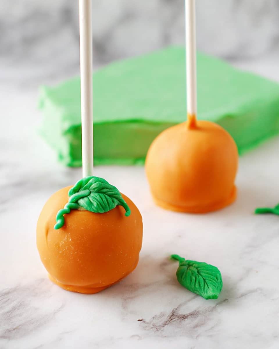 The image shows two round cake pops covered in smooth orange coating, each with a white stick inserted at the top center. On one cake pop, there is a small twisted green decoration that looks like a leaf and stem, placed near the top. Both cake pops sit on a white marbled surface with a square piece of green fondant in the background. Two small green decorations shaped like leaves and stems lie on the surface in front of the cake pops. photo taken with an iphone --ar 4:5 --v 7