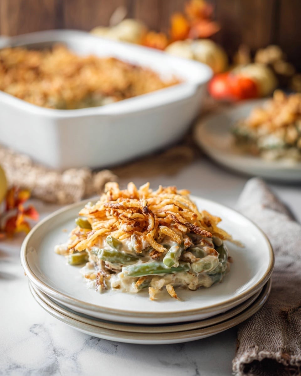 A creamy casserole with two visible layers sits on a stack of two white plates on a white marbled surface; the bottom layer is a pale beige creamy mixture with green beans mixed inside, topped by a crunchy golden-brown layer of fried onion strips. In the background, there is a large white baking dish filled with more of the casserole, showing the creamy beige and green mix beneath the golden topping, with part of a woman’s hand holding the plates. The scene is cozy with dim natural light and some blurred autumn-themed decorations in the back. Photo taken with an iphone --ar 4:5 --v 7