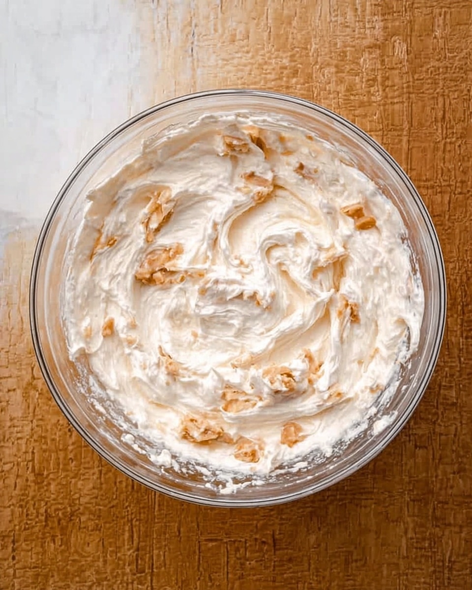 A clear glass bowl sits on a white marbled surface, filled with a creamy mixture that is off-white with a slightly uneven and soft texture. Small chunks of light brown pieces are scattered throughout the creamy base, giving the mix a bit of contrast and texture. The mixture looks thick but smooth, with swirls showing some movement and blending. photo taken with an iphone --ar 4:5 --v 7