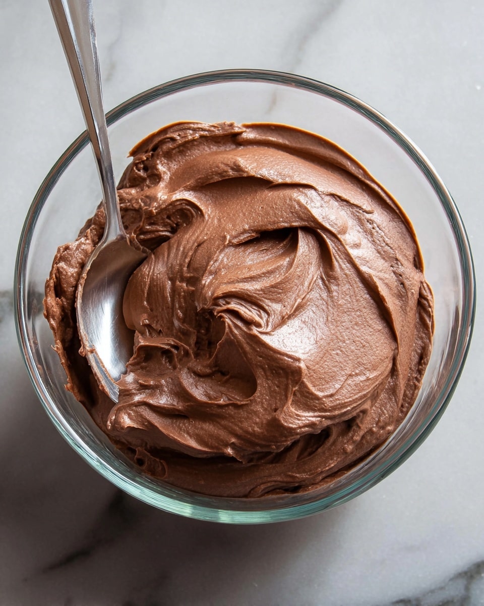 A clear glass bowl filled with a smooth, light brown chocolate mousse that has soft peaks and a creamy texture, with a silver spoon partially dipped into it on the left side of the bowl. The background shows a white marbled texture surface with two small red raspberries placed near the bowl at the back right. The lighting highlights the glossy surface of the mousse and the transparent edges of the glass bowl. Photo taken with an iphone --ar 4:5 --v 7