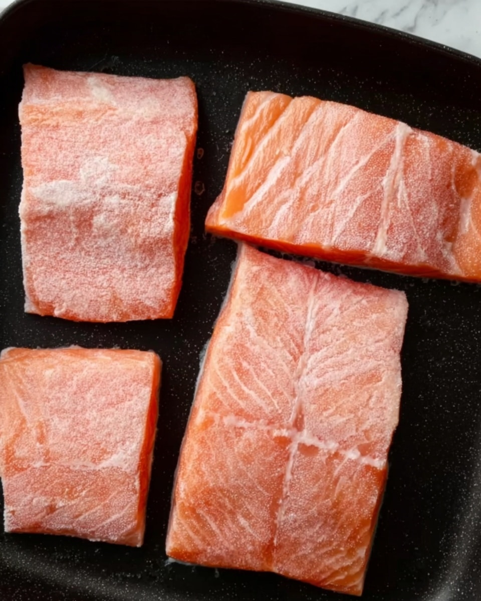 The image shows four raw salmon fillets with light pink skin and a slightly frosted surface, arranged in a black pan. Each fillet has a soft, smooth texture with faint white lines. The fillets are positioned close to each other but not overlapping, with the two larger ones on the top and bottom right and the smaller two in the middle left and bottom left. The background has a white marbled texture visible around the edges of the pan. Photo taken with an iphone --ar 4:5 --v 7