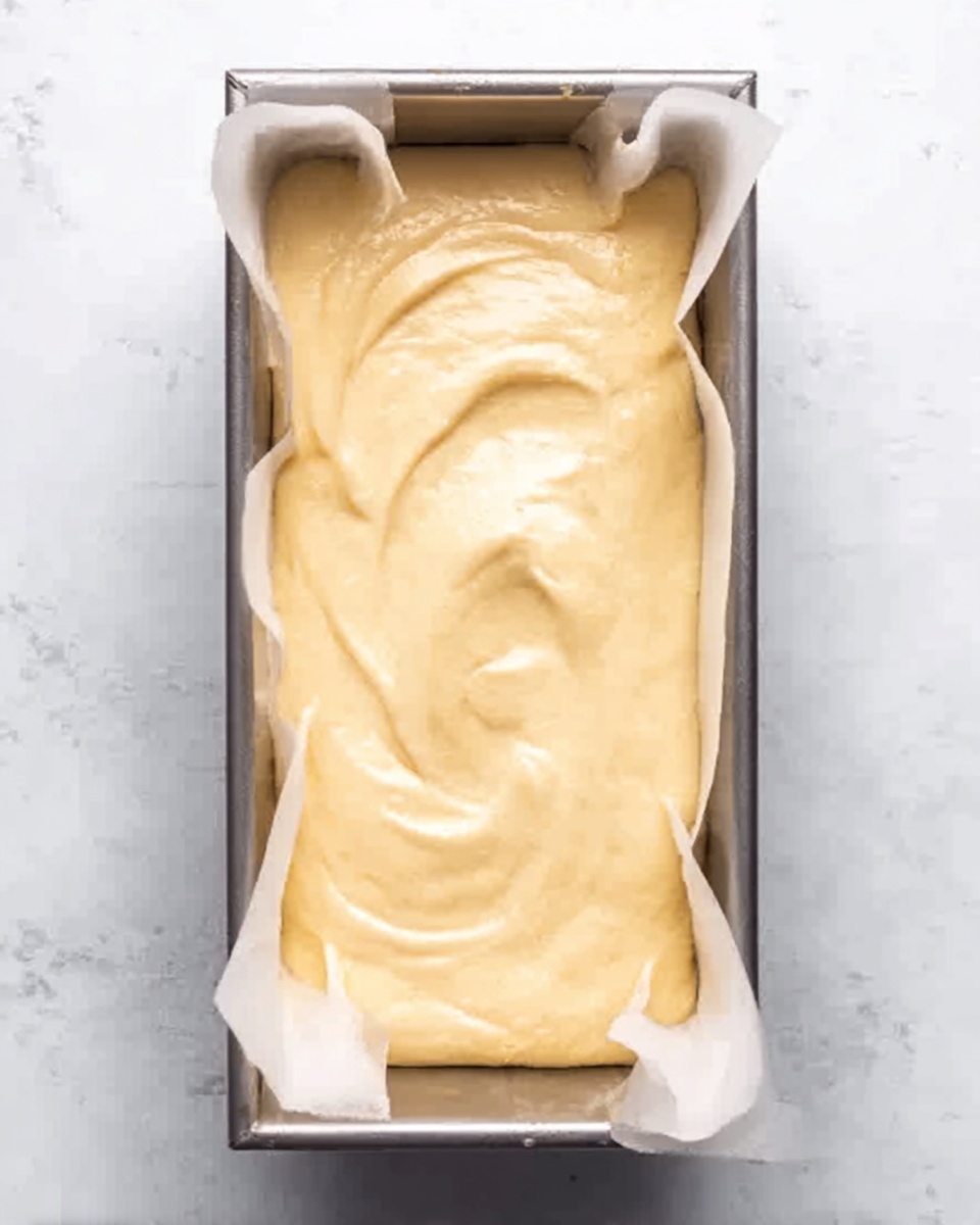 A rectangular silver baking pan lined with white paper holds a thick, creamy, pale yellow batter spread evenly inside. The batter looks smooth with gentle swirls and peaks on the surface, filling the pan almost to the top edges. The background is a white marbled texture. photo taken with an iphone --ar 4:5 --v 7