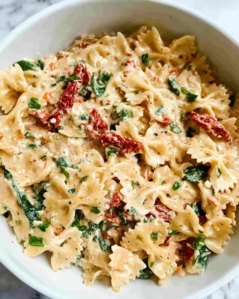 The image shows a white bowl filled with farfalle pasta covered in a creamy sauce. The pasta pieces are light beige with a soft, smooth texture. Mixed in are small pieces of red bell pepper and green spinach leaves, giving pops of color throughout. On top, a small green basil leaf is placed as garnish. The background features a white marbled surface. Photo taken with an iphone --ar 4:5 --v 7