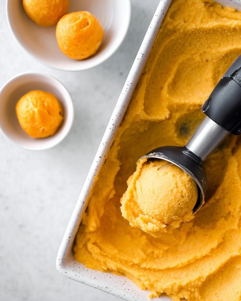 Two round scoops of bright yellow orange sorbet with a smooth and slightly shiny texture sit inside a white bowl. The sorbet has soft, creamy swirls and small air bubbles visible on the surface. Another similar bowl with more sorbet is blurred in the background on a white marbled surface. The light highlights the creamy texture and rich color of the sorbet in both bowls. photo taken with an iphone --ar 4:5 --v 7