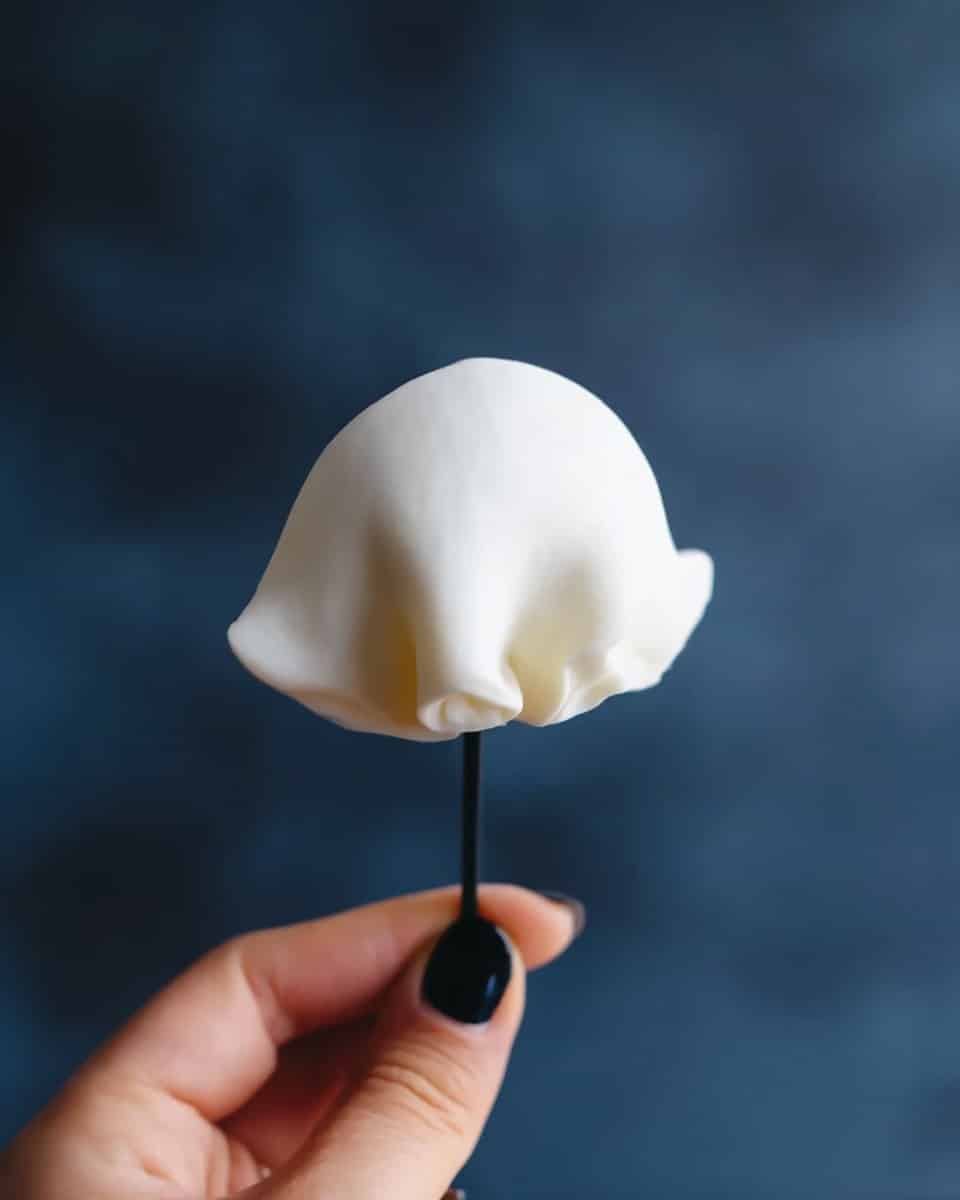 A close-up image shows a woman's hand with black nail polish holding a thin, white, dome-shaped piece of dough or fondant on a black stick. The background is blurry and dark blue, creating contrast with the white dough. The texture of the dough looks smooth and soft, slightly wrinkled at the bottom edges where it is pinched together. Photo taken with an iphone --ar 4:5 --v 7