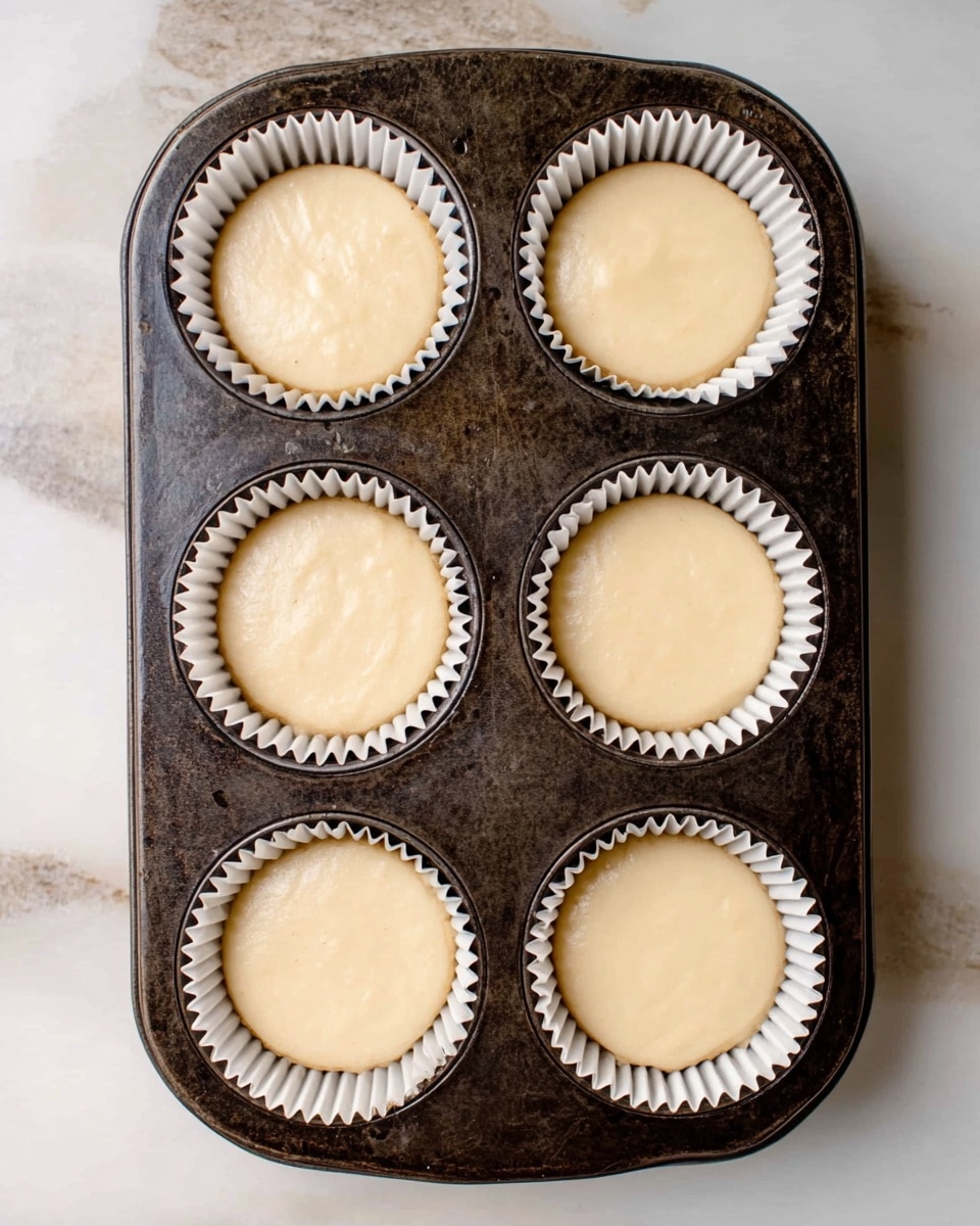 A dark metal muffin tray holds six cupcake liners, each filled with smooth, light beige batter that fills them nearly to the top. The liners are white with a ruffled edge, sitting evenly spaced in the tray. The tray rests on a white marbled surface that has soft grey veins running through it, giving a clean and bright background to the image. photo taken with an iphone --ar 4:5 --v 7