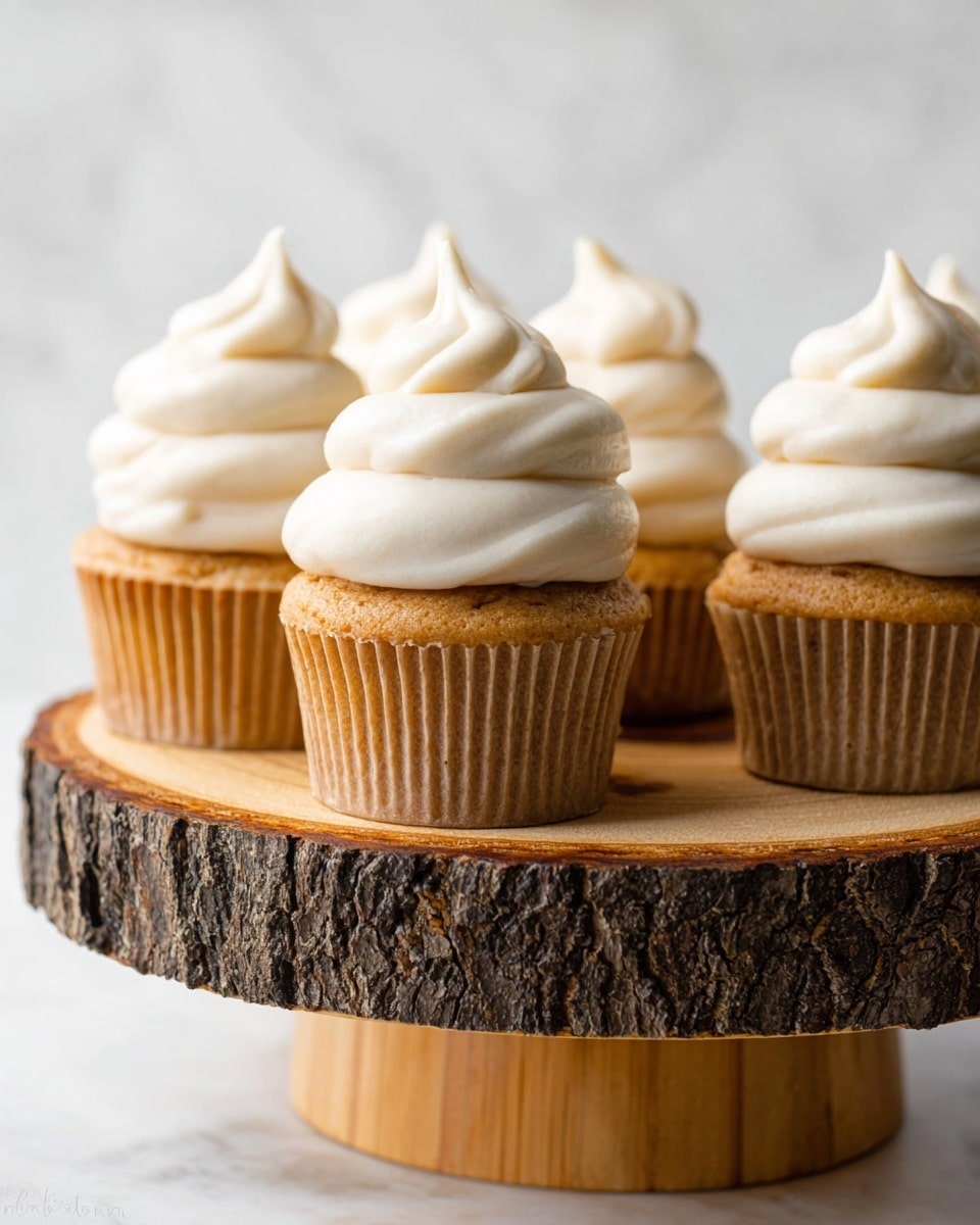 There are five cupcakes on a round wooden cake stand with a bark edge. Each cupcake has one layer of golden brown cake in a light beige paper cup. On top of each cake is a tall swirl of smooth white frosting that ends in a soft peak. The background is a light, white marbled texture, making the warm colors of the cupcakes and stand stand out. photo taken with an iphone --ar 4:5 --v 7