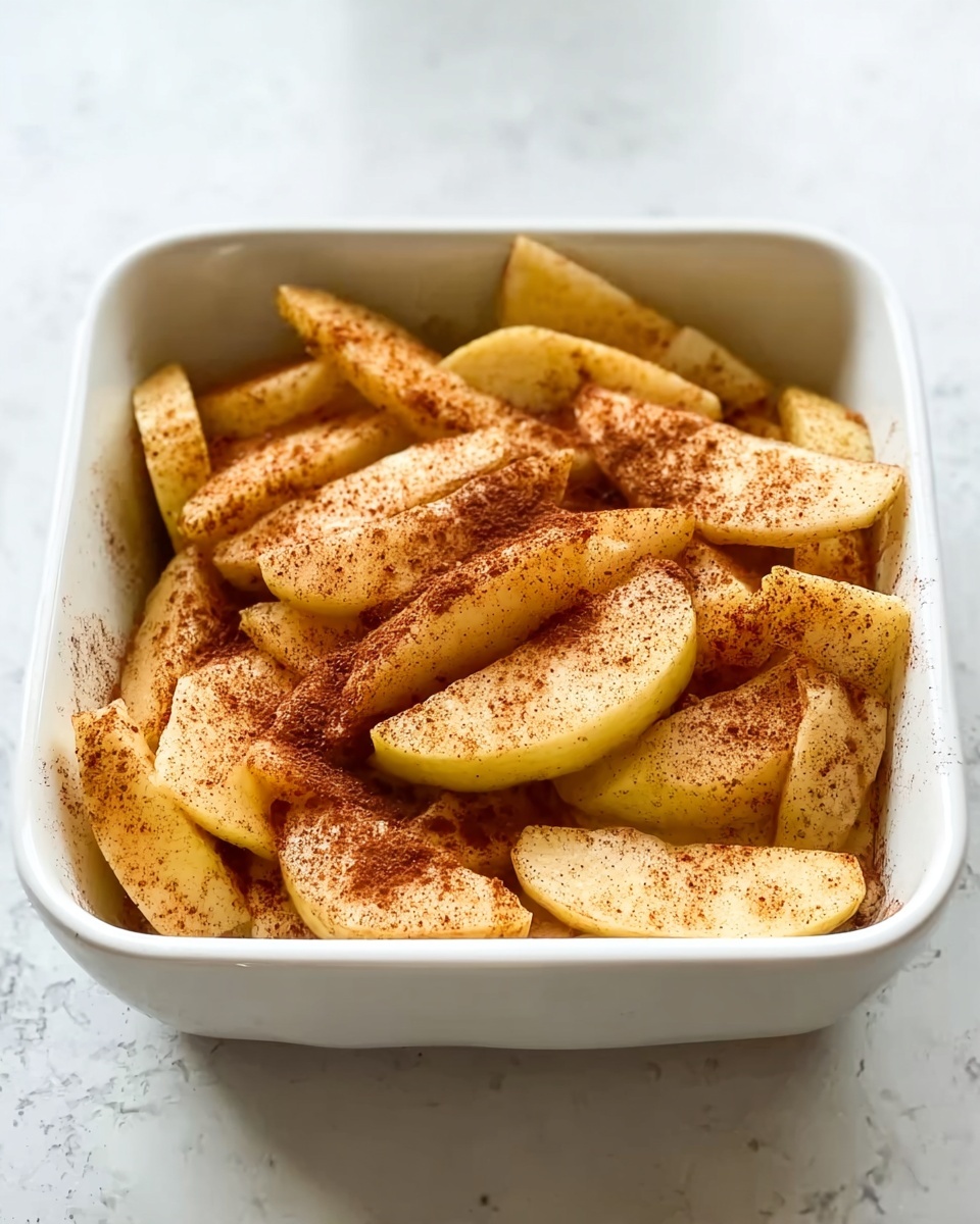 The image shows a white square dish filled with thick apple slices that are sprinkled evenly with cinnamon powder, giving the slices a warm brown color on top. The apple slices are stacked in a single layer, slightly overlapping each other, showing their light yellow flesh underneath the cinnamon. The dish sits on a white marbled surface, creating a clean and bright background. The photo was taken with an iphone --ar 4:5 --v 7