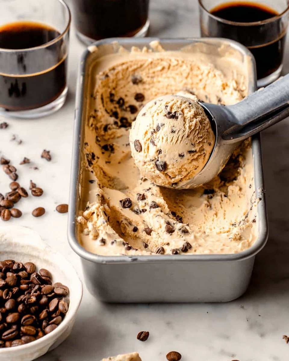 The image shows a metal container filled with light brown ice cream mixed with dark chocolate chips. A silver ice cream scoop is lifting a round scoop of ice cream from the container, with chocolate chips visible on the surface of the scoop. Next to the container, there is a white bowl filled with dark coffee beans and some scattered on the white marbled surface. In the background, two glasses with a dark liquid are partially visible. The whole scene is set on a white marbled texture, and the light highlights the creamy texture of the ice cream and the smooth metal of the scoop. Photo taken with an iphone --ar 4:5 --v 7