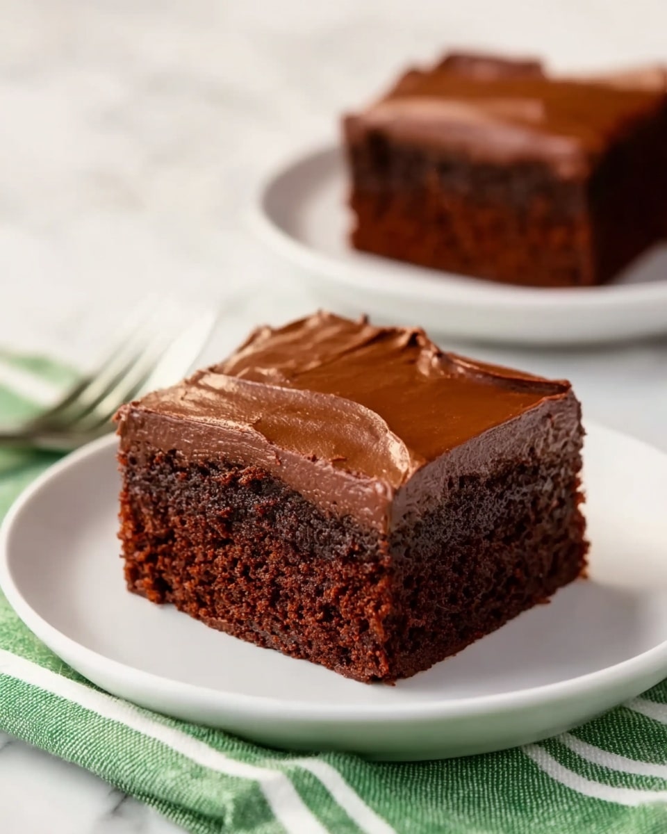 The image shows a thick, rectangular piece of chocolate cake placed on a white plate. The cake has two clear layers: the bottom layer is dark brown with a moist and soft texture, and the top layer is a smooth, glossy chocolate frosting with a rich, even spread. The cake sits on top of a green and white striped cloth napkin. In the background, there is another similar cake piece on a white plate, slightly out of focus. The surface beneath the plates is a white marbled texture. Photo taken with an iphone --ar 4:5 --v 7