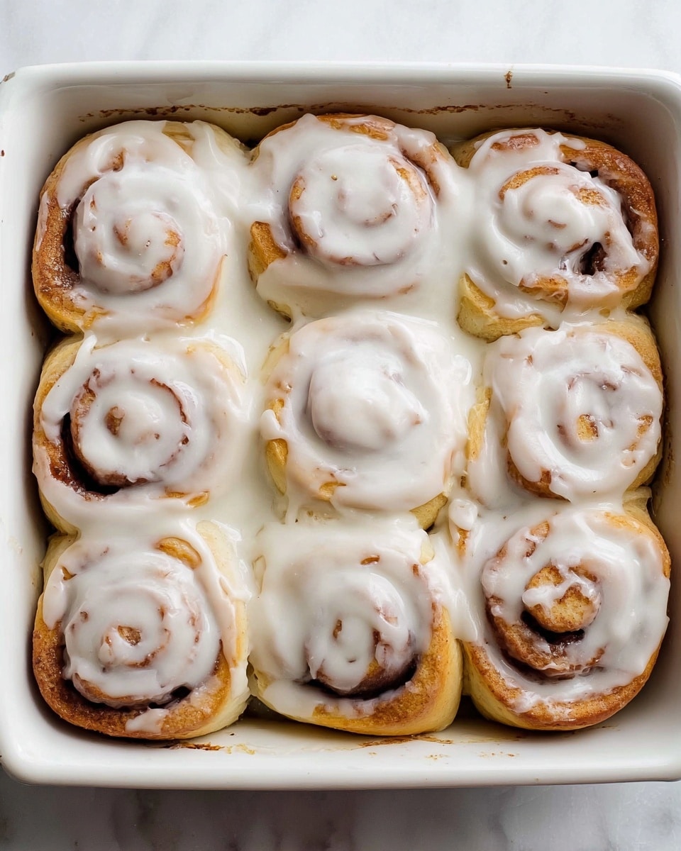 Nine cinnamon rolls are placed closely together in a white square baking dish, each roll showing soft, light brown dough with swirls of cinnamon inside. They are covered with a thick layer of white icing that spreads unevenly over the tops, allowing some of the cinnamon spiral patterns to show through. The rolls fill the dish almost edge to edge, and a small amount of the icing pools slightly in the empty spaces between them. The dish sits on a white marbled surface. photo taken with an iphone --ar 4:5 --v 7