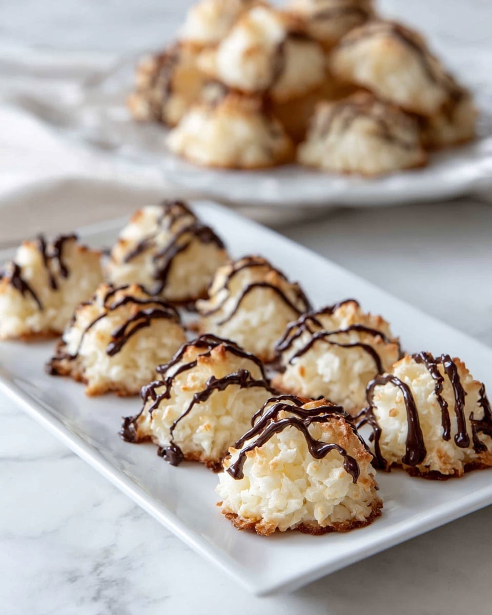 The image shows a white rectangular plate with two rows of small, rounded coconut macaroon cookies. Each cookie has a rough, textured surface with a golden brown tint on the edges and a creamy white center. They are drizzled with thin lines of dark chocolate in an irregular pattern, adding contrast to the light cookies. In the background, there is another white plate with more macaroons slightly out of focus, all set on a white marbled surface. photo taken with an iphone --ar 4:5 --v 7