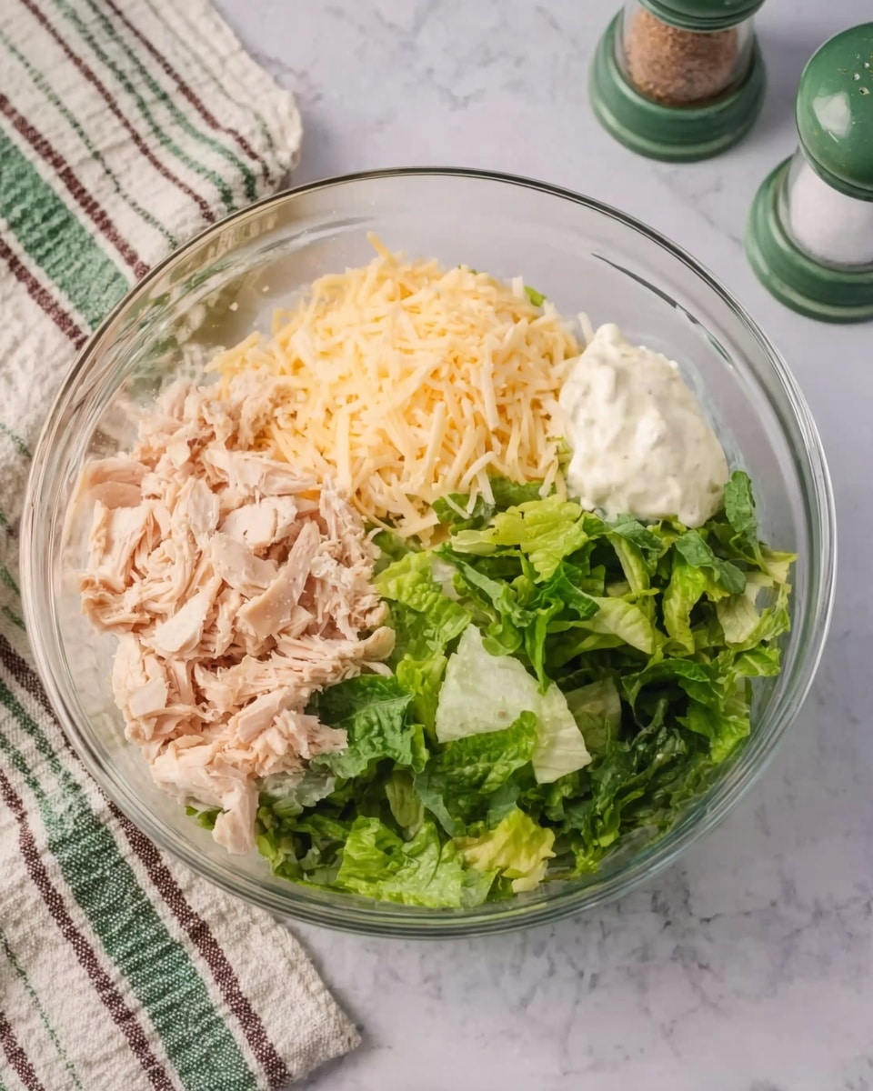 A clear glass bowl sits on a white marbled surface, filled with four distinct sections of ingredients. On the left, there are light pink shredded pieces, likely cooked chicken. At the top, there is a mound of pale yellow shredded cheese. On the right side next to the cheese, a dollop of white creamy dressing rests. The bottom section contains fresh, bright green leafy lettuce. The bowl is placed near a patterned cloth napkin with green, white, and brown stripes, and salt and pepper shakers with green tops are visible in the background. photo taken with an iphone --ar 4:5 --v 7
