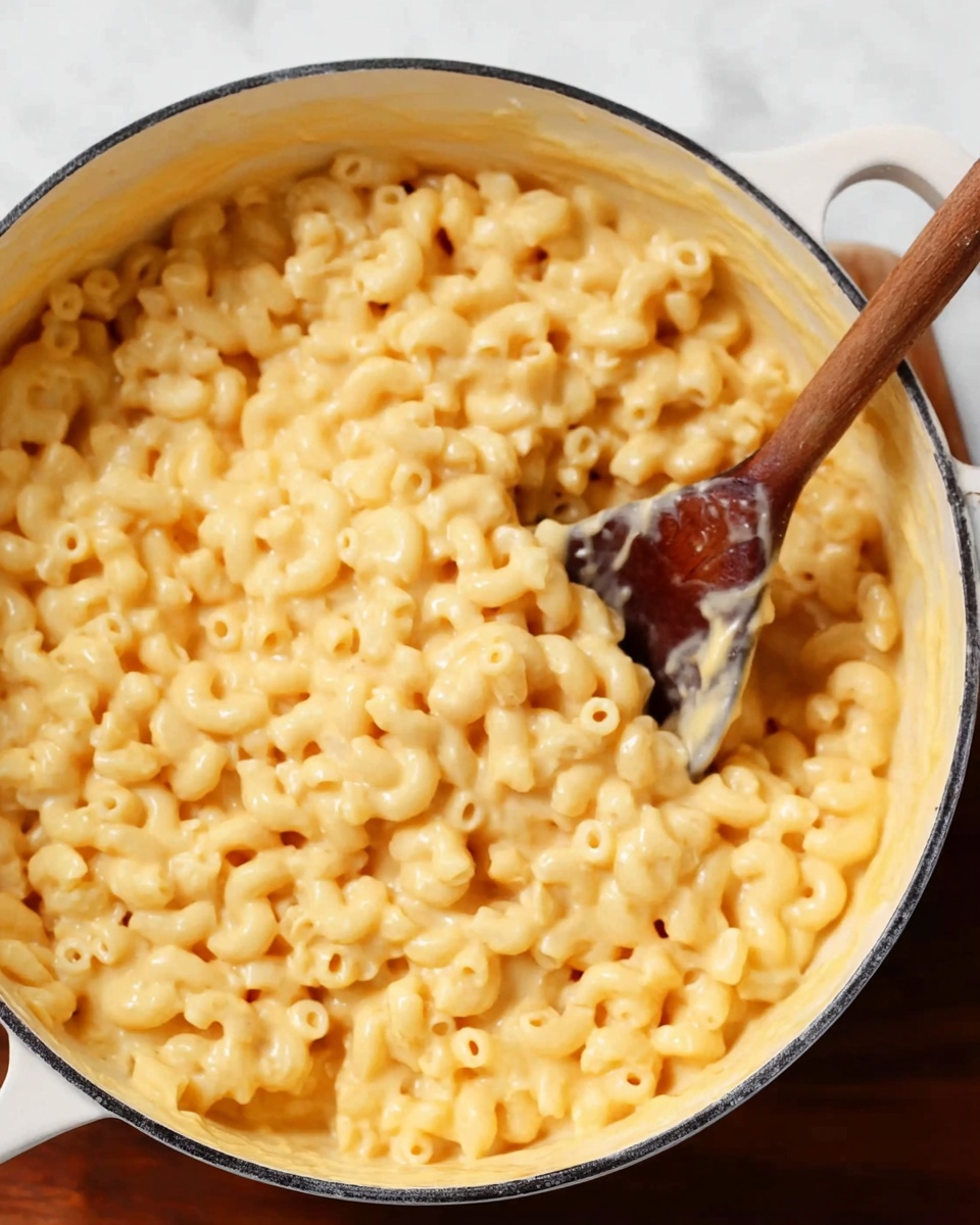 A close-up view of a white round pot filled with creamy macaroni and cheese. The macaroni pasta is small and curved, covered evenly with smooth, light yellow cheese sauce. A wooden spoon is partly dipped into the pot, lifting a portion of the cheesy pasta, showing a thick and soft texture. The pot sits on a white marbled surface that contrasts with the warm colors of the food, adding brightness to the scene. Photo taken with an iphone --ar 4:5 --v 7