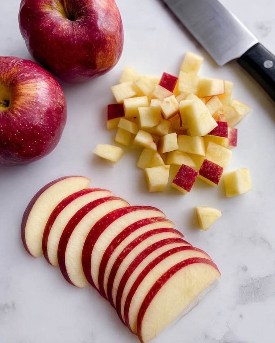 The image shows a white marbled surface with a group of apple slices and apple cubes placed near each other. At the bottom left, there are nine thin, fan-shaped apple slices with red skin and light yellow flesh, slightly overlapping each other in a neat row. Above and to the right of the slices, there is a pile of small apple cubes with red edges and light yellow centers, arranged loosely in a small mound. In the top right corner, part of a large kitchen knife with a black handle and shiny blade rests on the surface. In the top left corner, two whole red apples with a shiny, smooth skin are placed close together. Photo taken with an iphone --ar 4:5 --v 7
