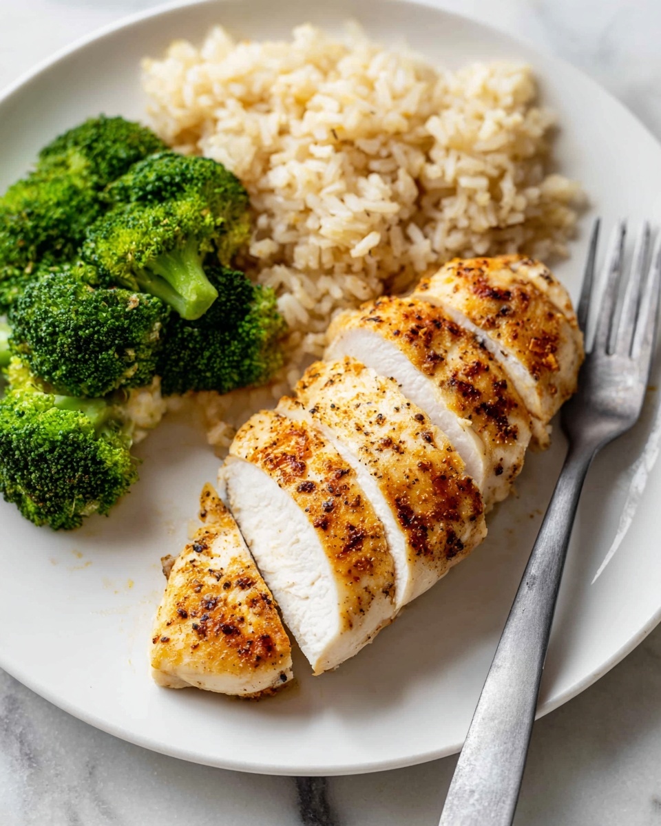 A white plate holds a meal with three parts. On the right, there is a cooked chicken piece, light golden brown with small black seasoning dots, sliced into thick sections, showing white moist inside. To the left of the chicken, a small pile of bright green steamed broccoli florets with textured tops is arranged. Above the chicken, off to the right, there is a serving of soft, light beige brown rice with separated grains. The plate sits on a white marbled surface, and a long gray spoon rests near the chicken. photo taken with an iphone --ar 4:5 --v 7