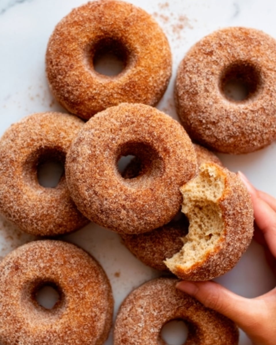 A stack of three cinnamon sugar donuts sits on a white ceramic plate with a brown decorative edge. The donuts are golden brown and covered in a rough layer of sugar and cinnamon, giving them a grainy texture. The top donut has a bite taken out of it, revealing a soft, light, and fluffy inside. The plate rests on a white marbled surface with a beige cloth underneath, and part of a soft pink cloth is visible on the side. In the background, there is a white cup filled with coffee and some blurred green leaves and a pink vase photo taken with an iphone --ar 4:5 --v 7