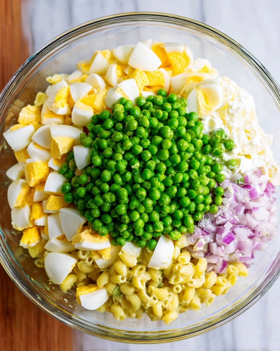 A clear glass bowl sits on a white marbled surface, filled with layers of colorful ingredients. At the bottom, pale yellow small pasta pieces form the first layer, topped by a thick layer of creamy white dressing mixed with finely chopped light purple onions on the right side. On the left side, there is a dense pile of chopped hard-boiled eggs showing bright yellow yolks and white edges. On top, a generous mound of vibrant green peas forms the final layer, sitting in the center. Photo taken with an iphone --ar 4:5 --v 7