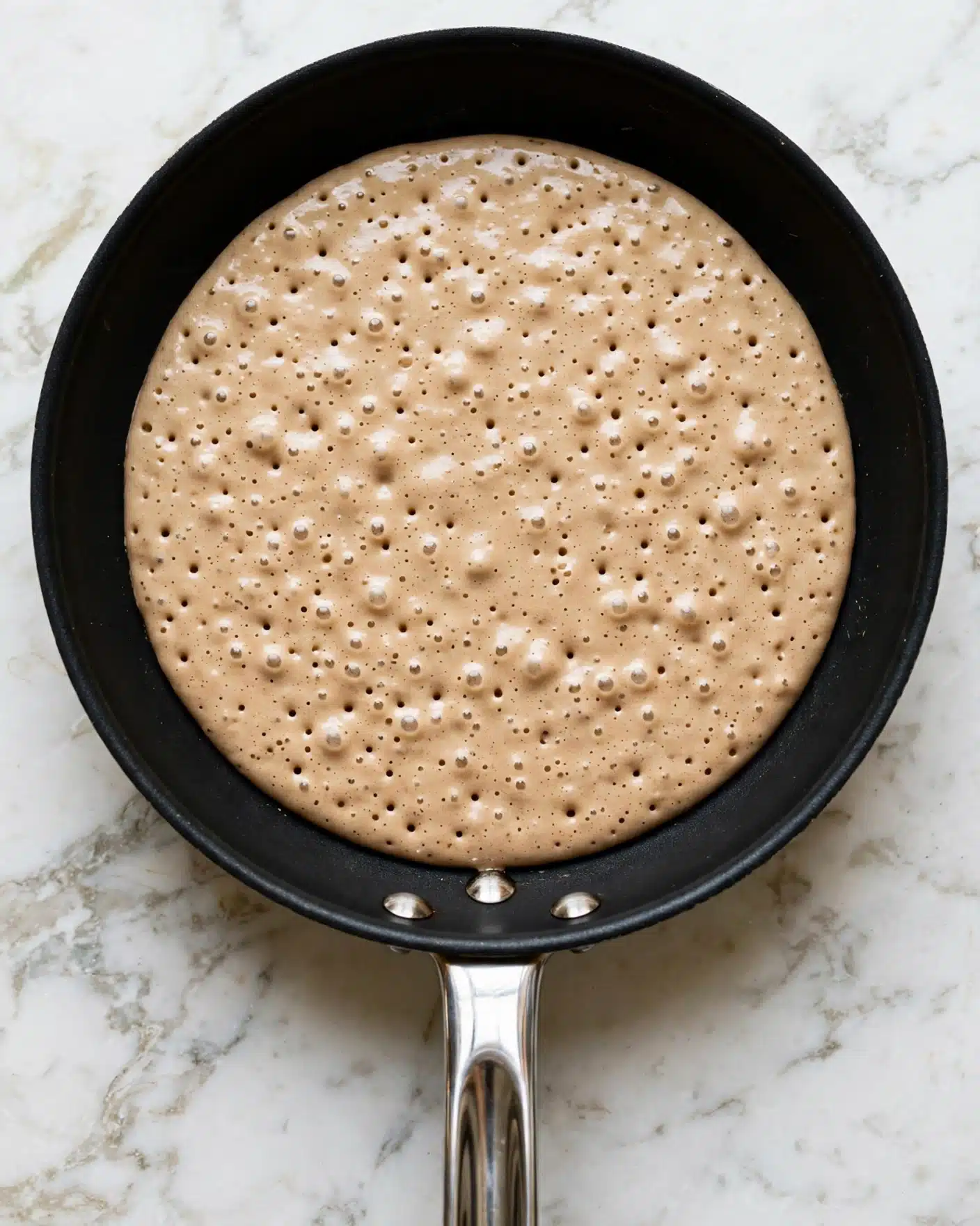 A single round pancake layer is cooking in a black frying pan, with a smooth light brown batter that shows small bubbles rising on the surface. The batter is evenly spread inside the pan, and the pan has a shiny metallic handle pointing towards the bottom of the image. The frying pan is placed on a white marbled surface. Photo taken with an iphone --ar 4:5 --v 7