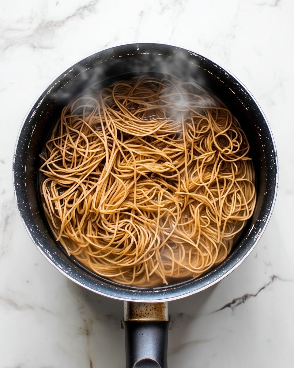 A top view of a black pot filled with light brown noodles cooking in water, the noodles loosely piled and partially submerged, with steam rising gently above the surface. The pot has a black handle with a slightly worn area near the base where it meets the pot. The pot is placed on a white marbled surface that shows faint grey and beige veining. The image is bright with natural light, making the textures of the noodles and pot clear. photo taken with an iphone --ar 4:5 --v 7