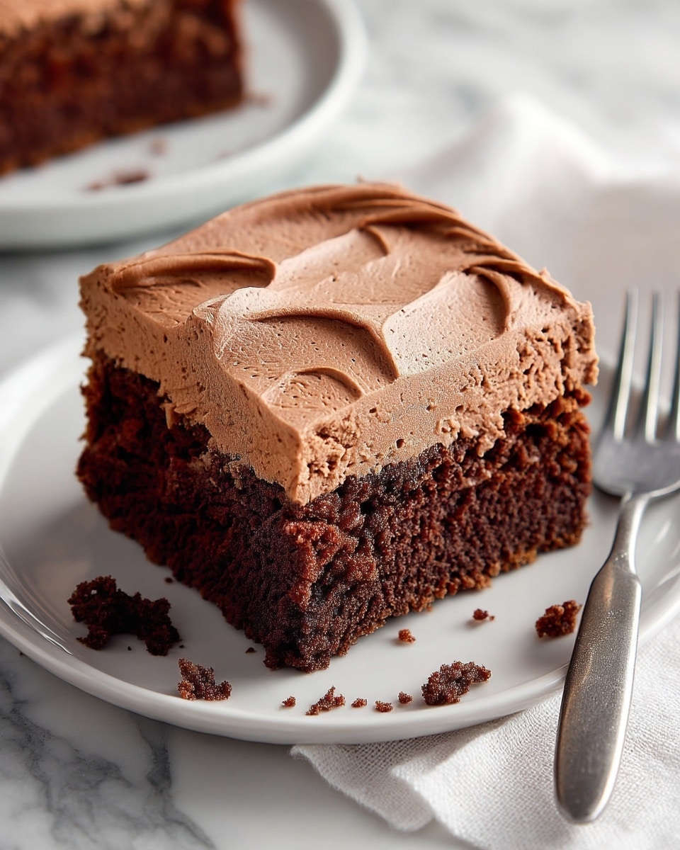 The image shows one square piece of chocolate cake on a white plate. The cake has two layers: the bottom layer is thick and dark brown with a soft, crumbly texture, while the top layer is a lighter brown, smooth, and creamy frosting spread evenly with swirled patterns. There are a few small crumbs scattered around the cake on the plate. The plate sits on a white marbled surface with a white cloth and the edge of a fork visible on the right. Photo taken with an iphone --ar 4:5 --v 7