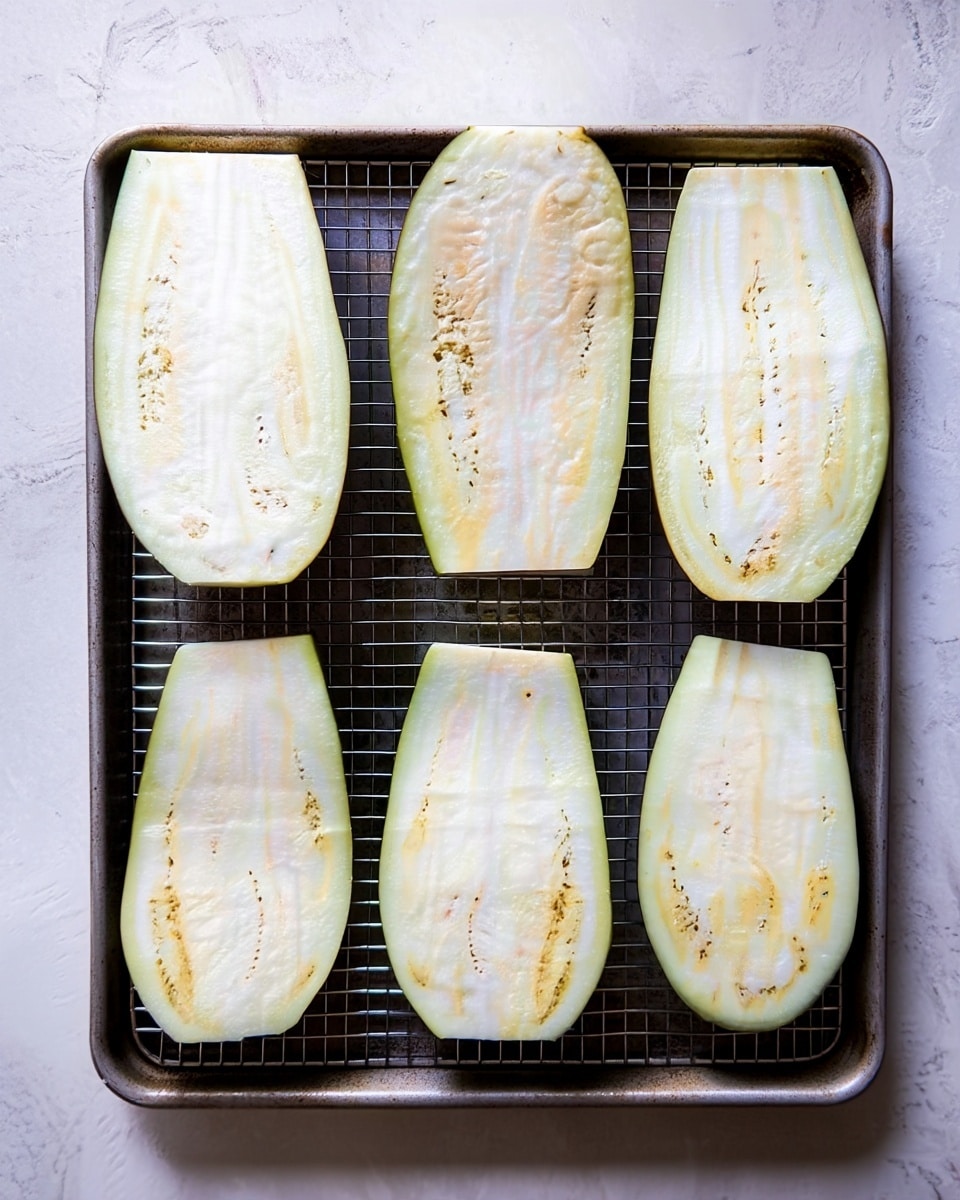 The image shows six long, oval slices of eggplant arranged in two rows of three on a metal wire rack placed inside a dark baking tray. Each eggplant slice has a pale creamy color with small, scattered light brown seeds visible inside. The eggplant skin is thin and light green, framing the pale interior. The tray sits on a white marbled surface. photo taken with an iphone --ar 4:5 --v 7