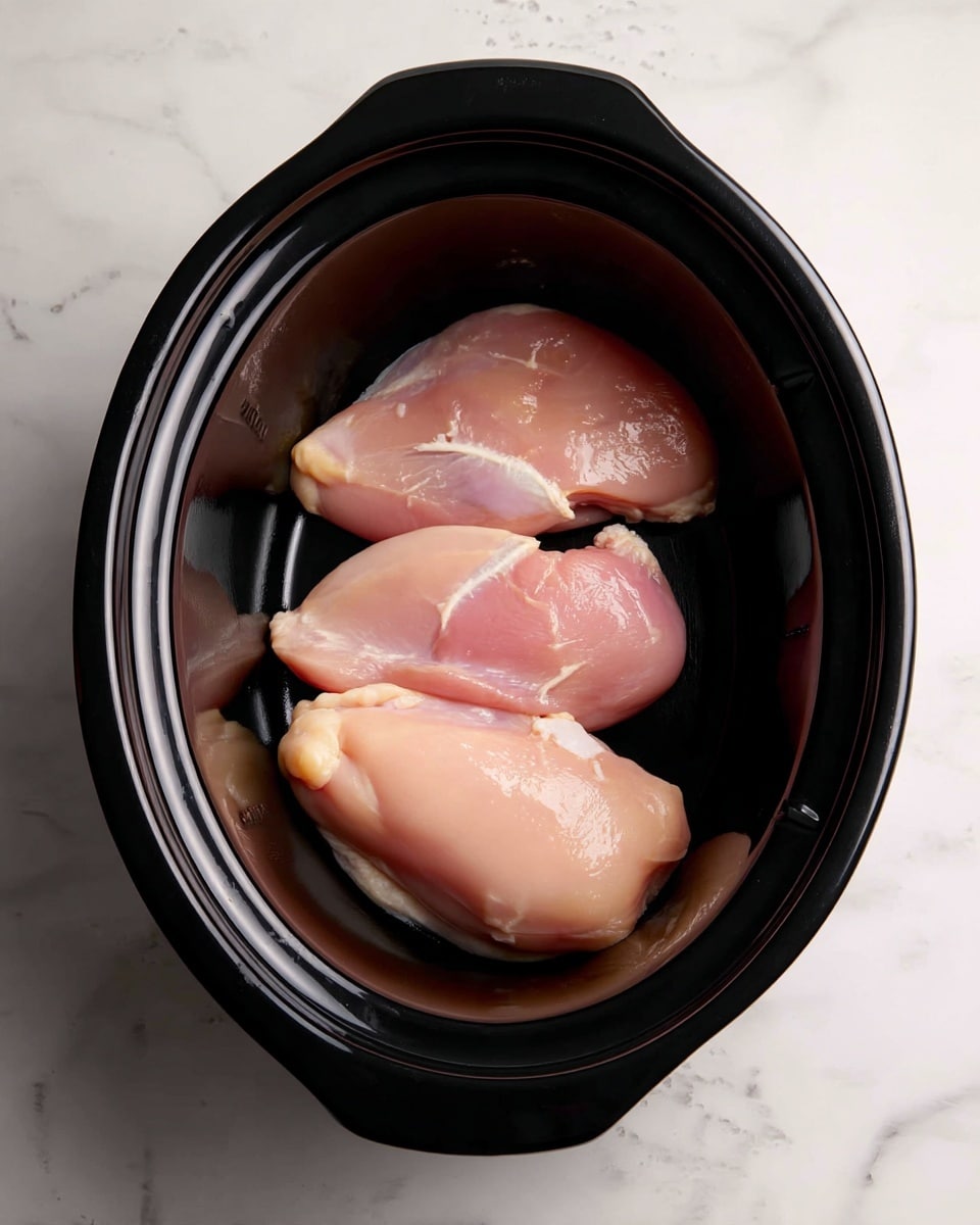 A black oval slow cooker with three raw pale pink chicken pieces inside, arranged close together on the bottom. The cooker is placed on a white marbled surface. The chicken pieces have smooth texture and light shine from the surface. Photo taken with an iphone --ar 4:5 --v 7