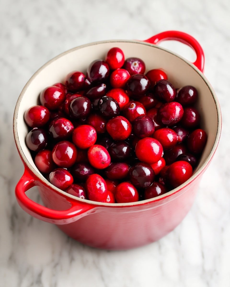 A white interior pot with red handles is filled with shiny, dark red cranberries, some falling into the pot. The cranberries are smooth and round, packed close together inside the pot. The pot sits on a stove, but the visible background is replaced by a white marbled texture. The bright red of the cranberries stands out against the clean white inside of the pot and the white marble surface photo taken with an iphone --ar 4:5 --v 7
