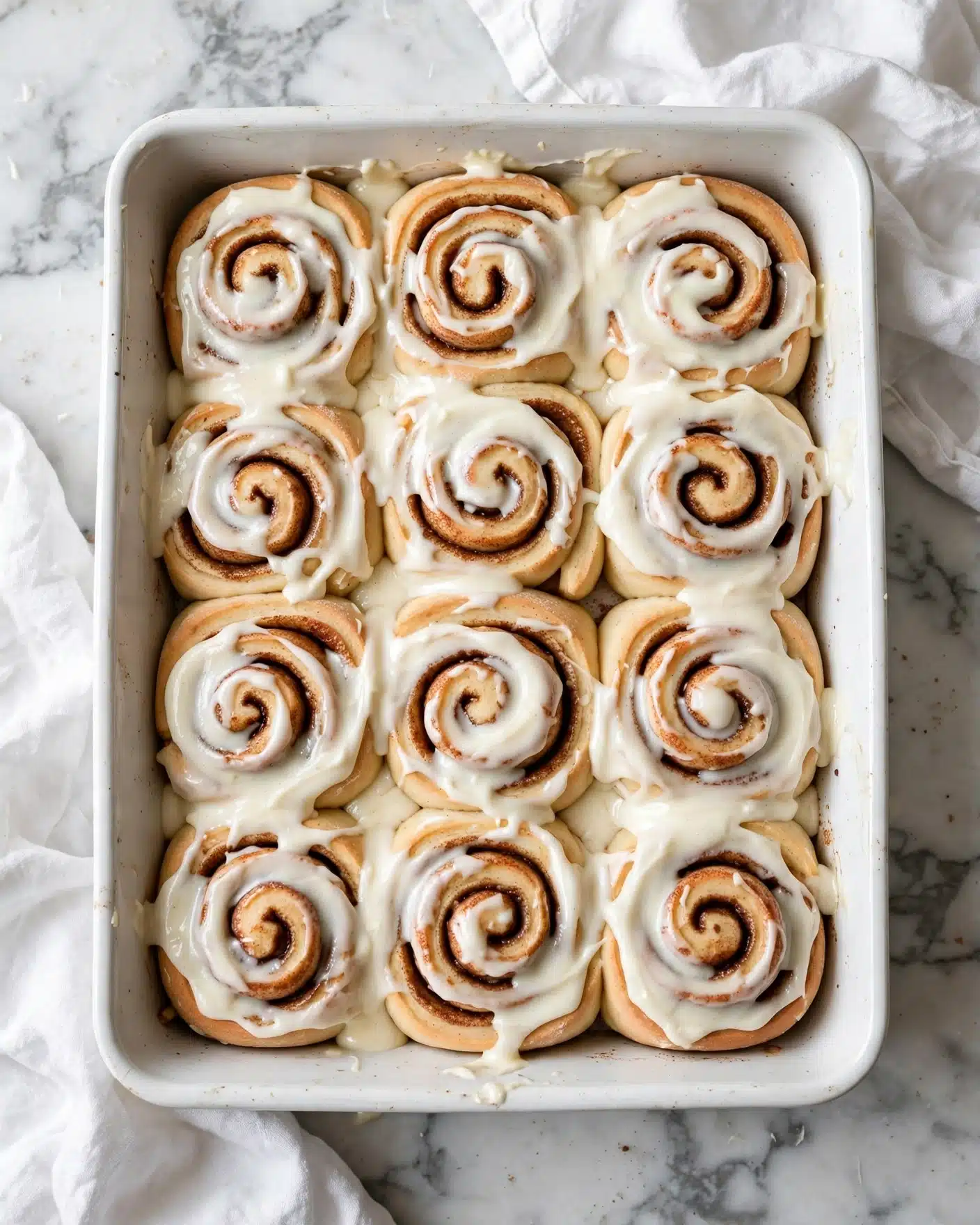 Soft and Fluffy Homemade Cinnamon Rolls Recipe 6 A white rectangular baking dish filled with twelve cinnamon rolls closely placed side by side in a 3 by 4 layout. Each roll is light golden brown with visible spirals of cinnamon filling inside, topped thickly with creamy white icing that spreads softly over the edges and some parts of the swirls. The dish is set on a white marbled surface with a crumpled white cloth partially visible around the edges. Photo taken with an iphone --ar 4:5 --v 7