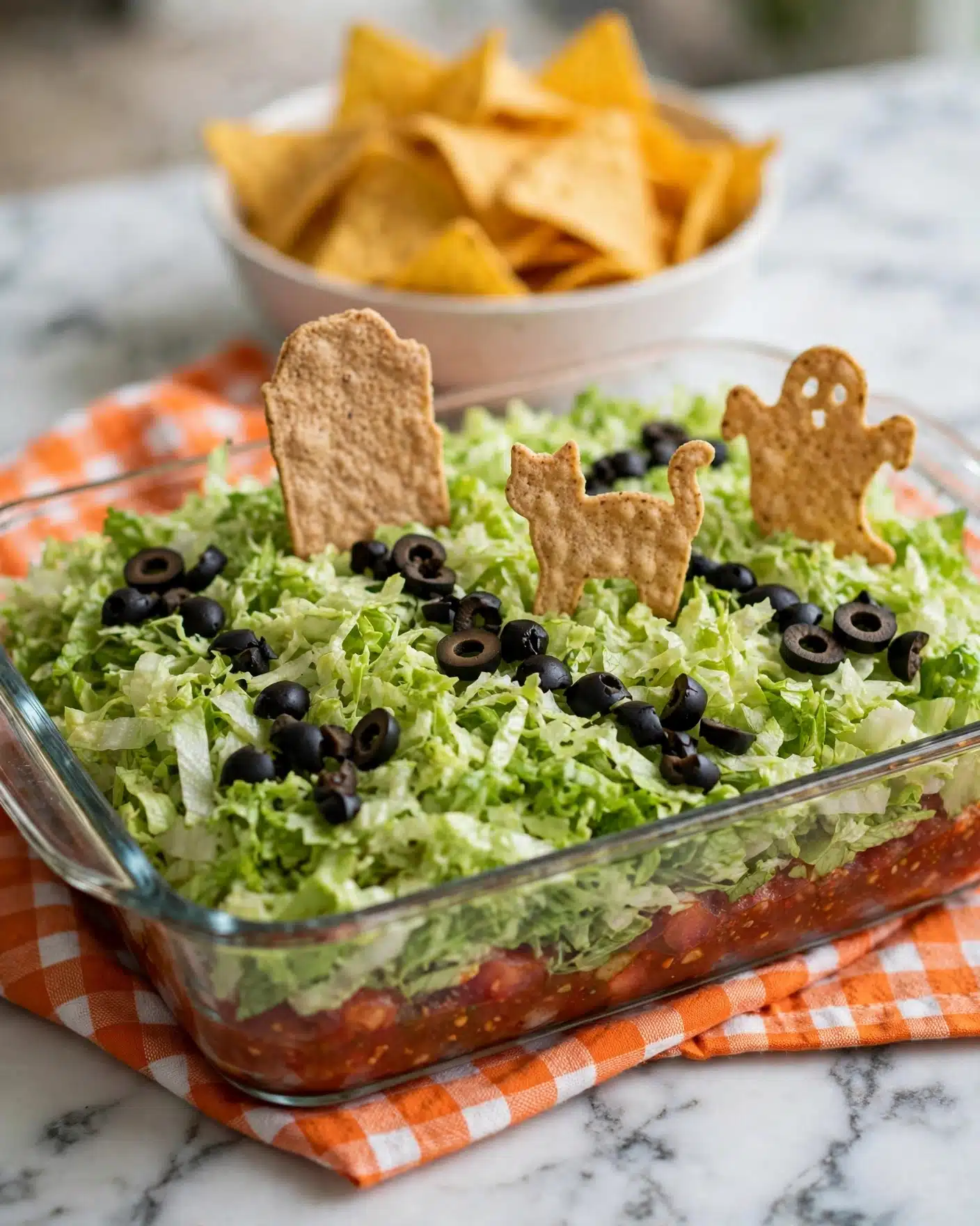 A clear glass rectangular dish filled with several layers including a reddish layer at the bottom mostly hidden, topped with a thick layer of light green shredded lettuce covering the whole dish. On top of the lettuce, there are black olive pieces scattered to form small paths and clusters. Standing upright in the lettuce are round and irregularly shaped light brown taco chips, some shaped like tombstones, a cat, and a ghost, arranged to look like a Halloween graveyard scene. The dish sits on an orange cloth with white checks on a white marbled surface. In the background, there is a white bowl filled with yellow-orange triangular chips. photo taken with an iphone --ar 4:5 --v 7
